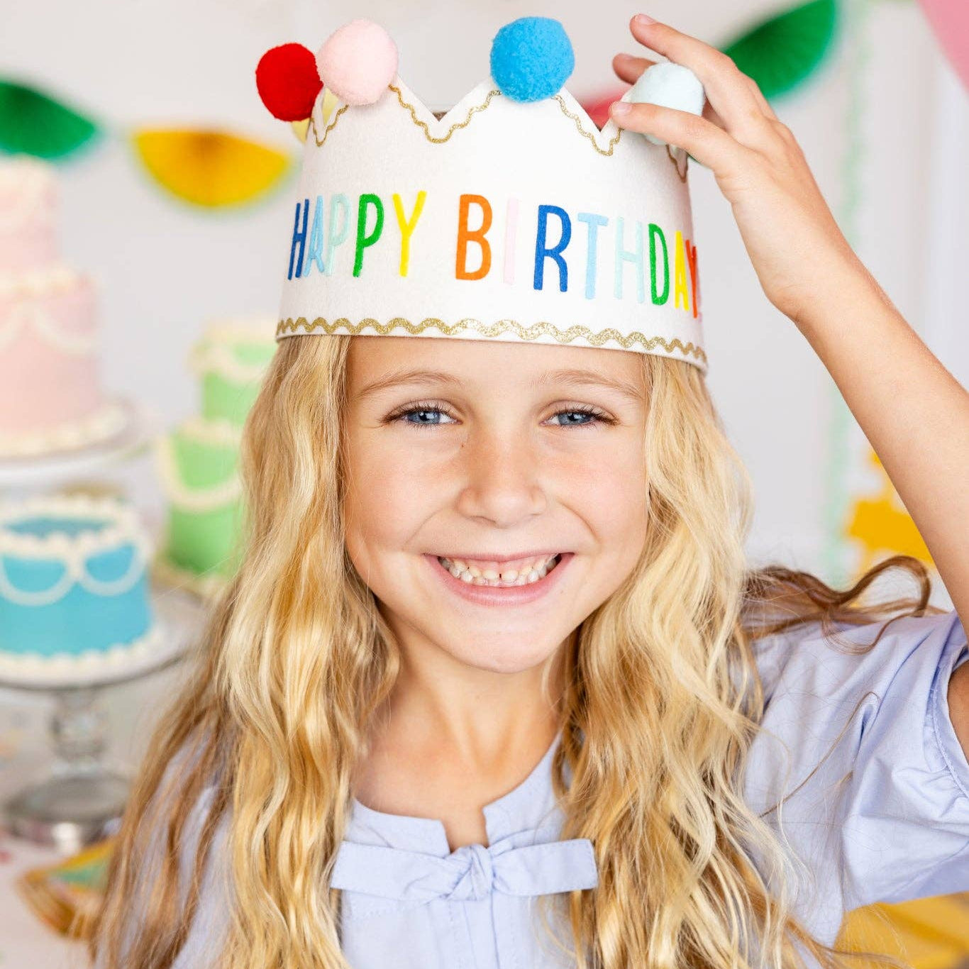 Child wearing a 'Happy Birthday' crown with colorful pom-poms, standing in front of a festive background.