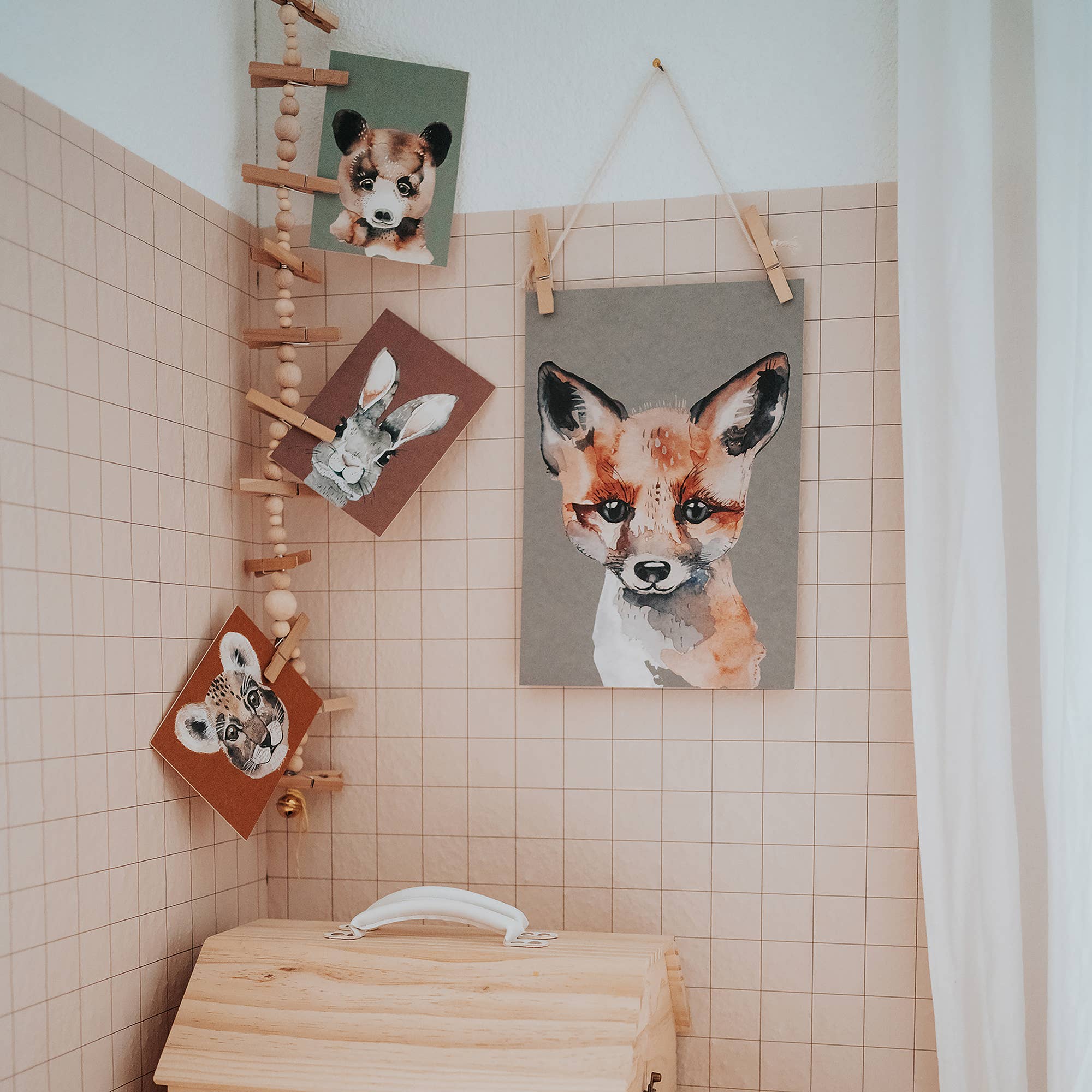 A styled interior corner showing a square canvas print of a fox head hanging on a tiled wall, alongside other animal portrait prints clipped to a wooden ladder shelf. A light wood chest sits below.