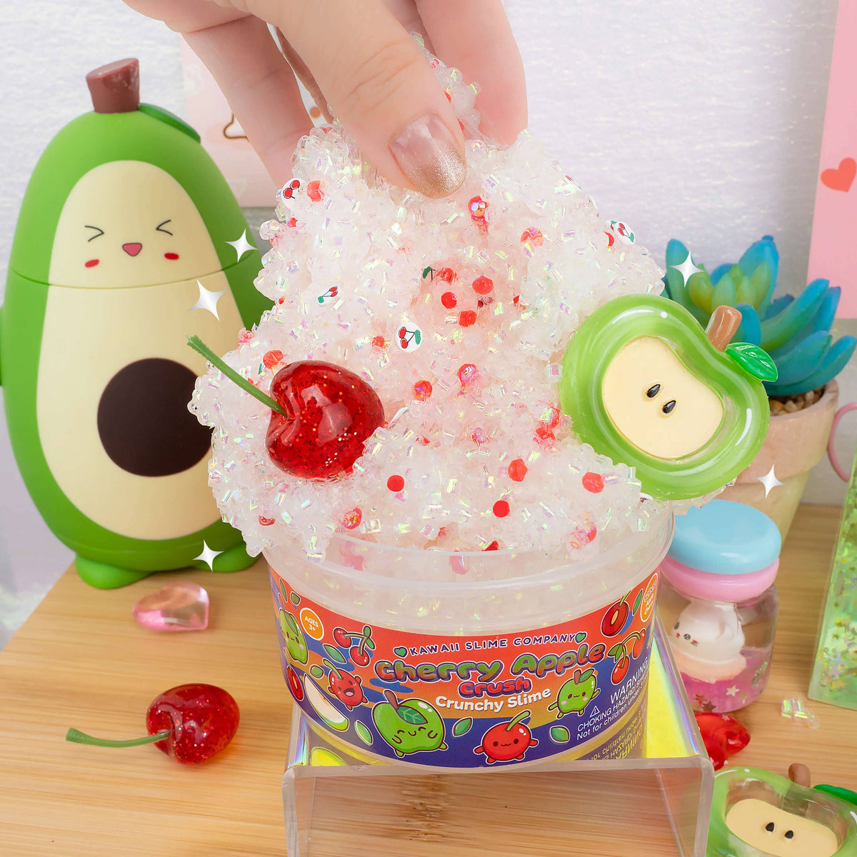 A hand is pressing down on a clear, glittery, whipped slime product in a plastic tub labeled 'Cherry Apple Crunchy Slime' by Pocket Candy Company. The white, translucent slime is decorated with red glitter and topped with a red cherry decoration and a green apple-shaped charm.