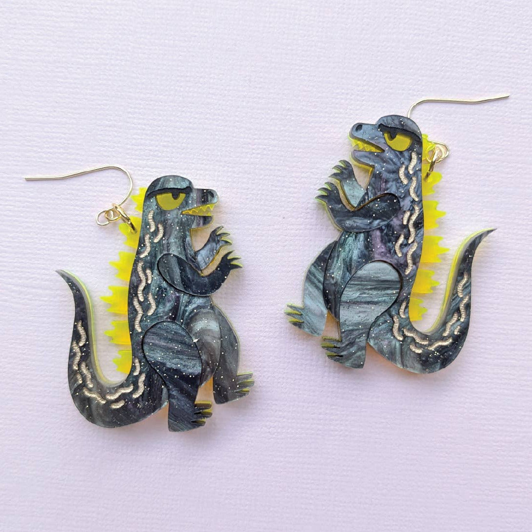 Dinosaur-shaped earrings on a light purple background with 'Sylvia Draws' branding.