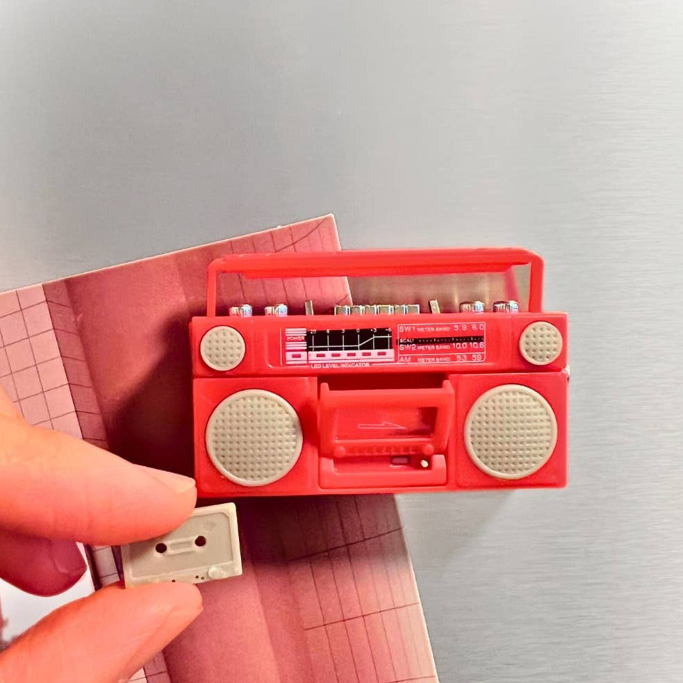 Red toy boombox with cassette tape held by a hand on a light gray background