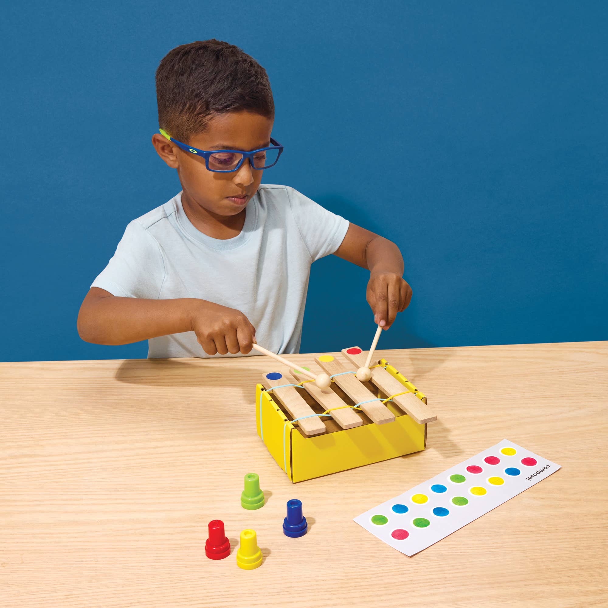A child wearing glasses sits at a light wood table playing a wooden xylophone toy with colorful striker rods and pegs, against a solid blue background.