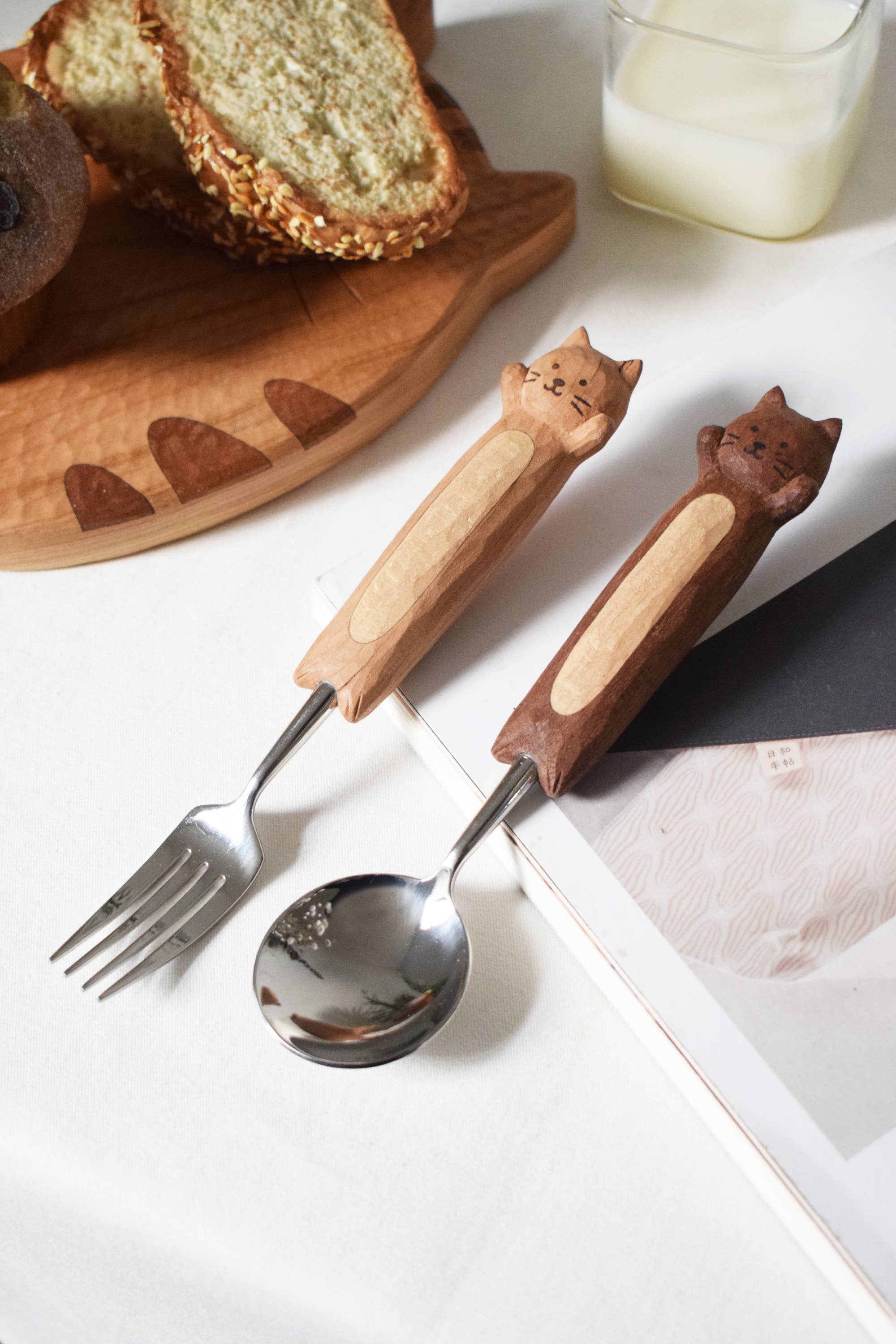 Cat-shaped wooden handles on a fork and spoon with a cake and glass of milk in the background.