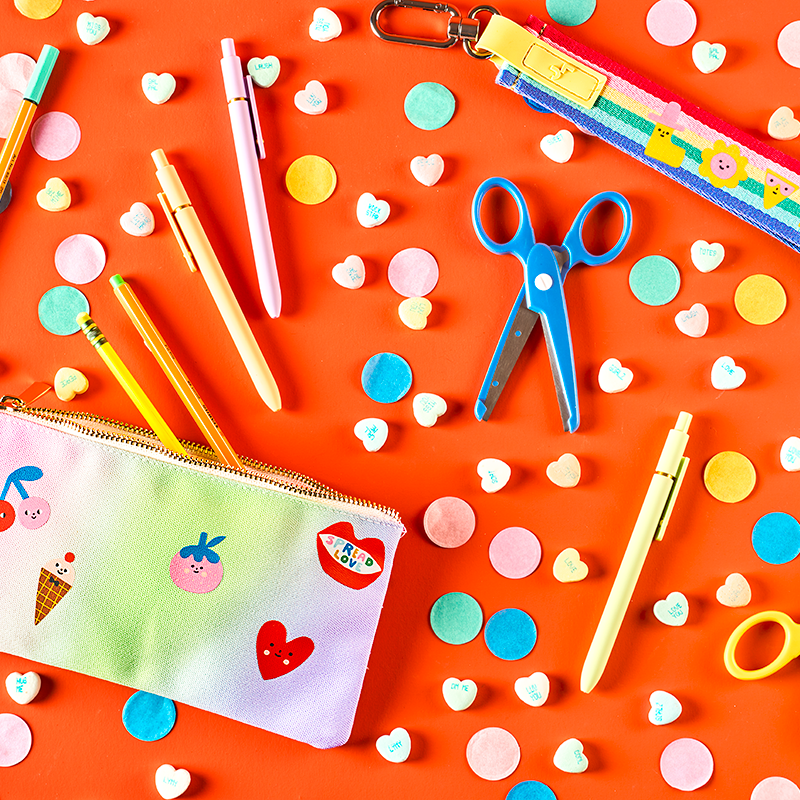 Colorful stationery items including pens, scissors, and a pouch on a bright orange background with polka dots.