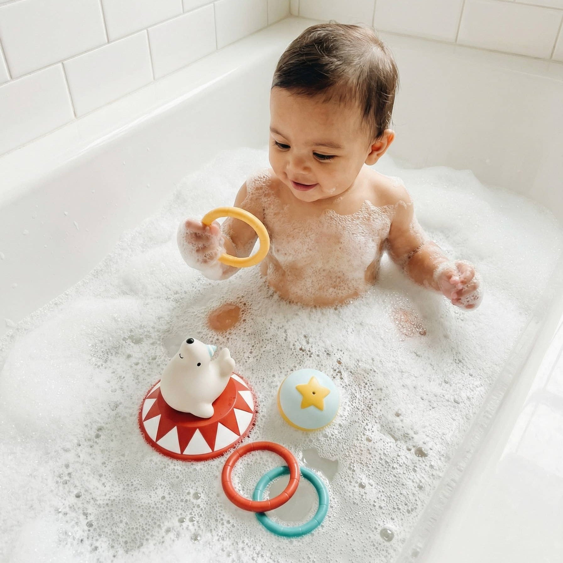 A baby sitting in a bubble-filled bathtub playing with colorful bath toys, including a yellow ring, a red and white shark toy, and a blue star toy.