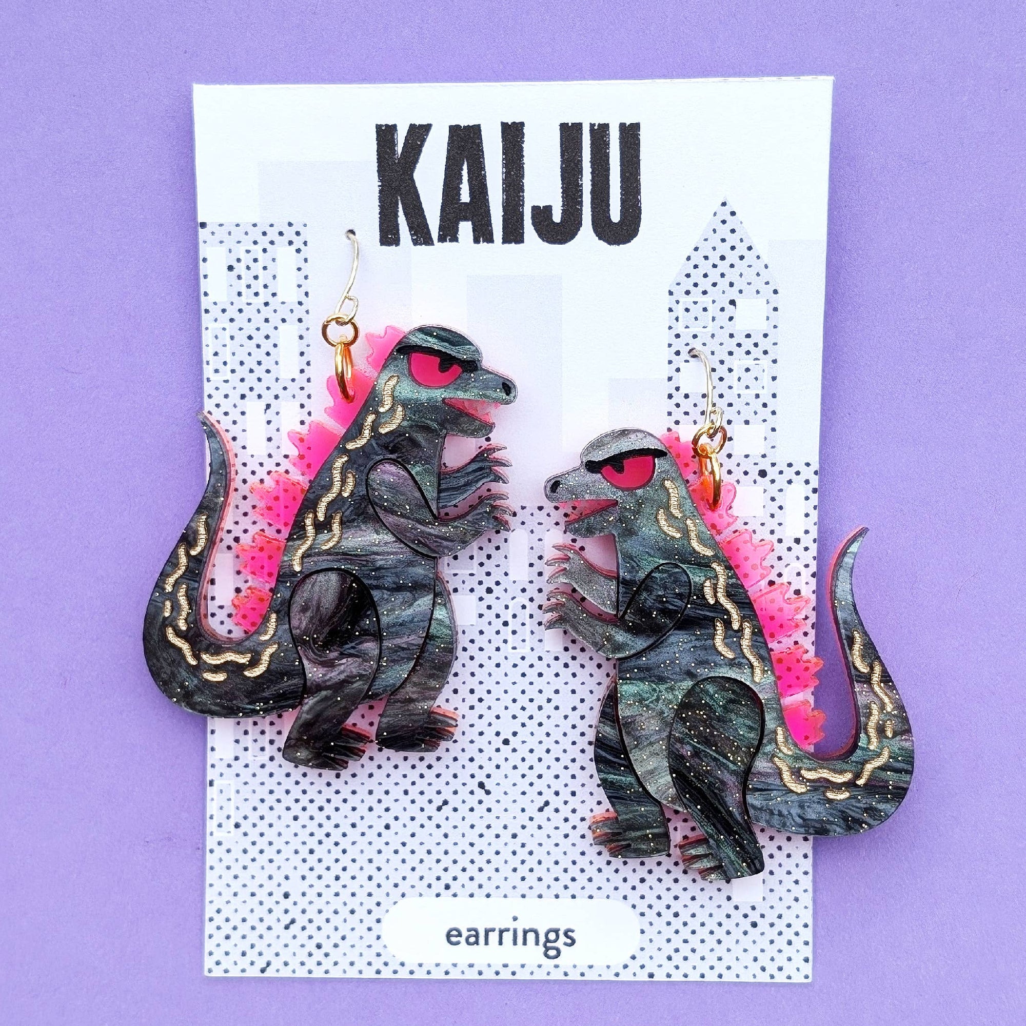 Kaiju earrings on a card with 'Sylvia Draws!' branding against a purple background