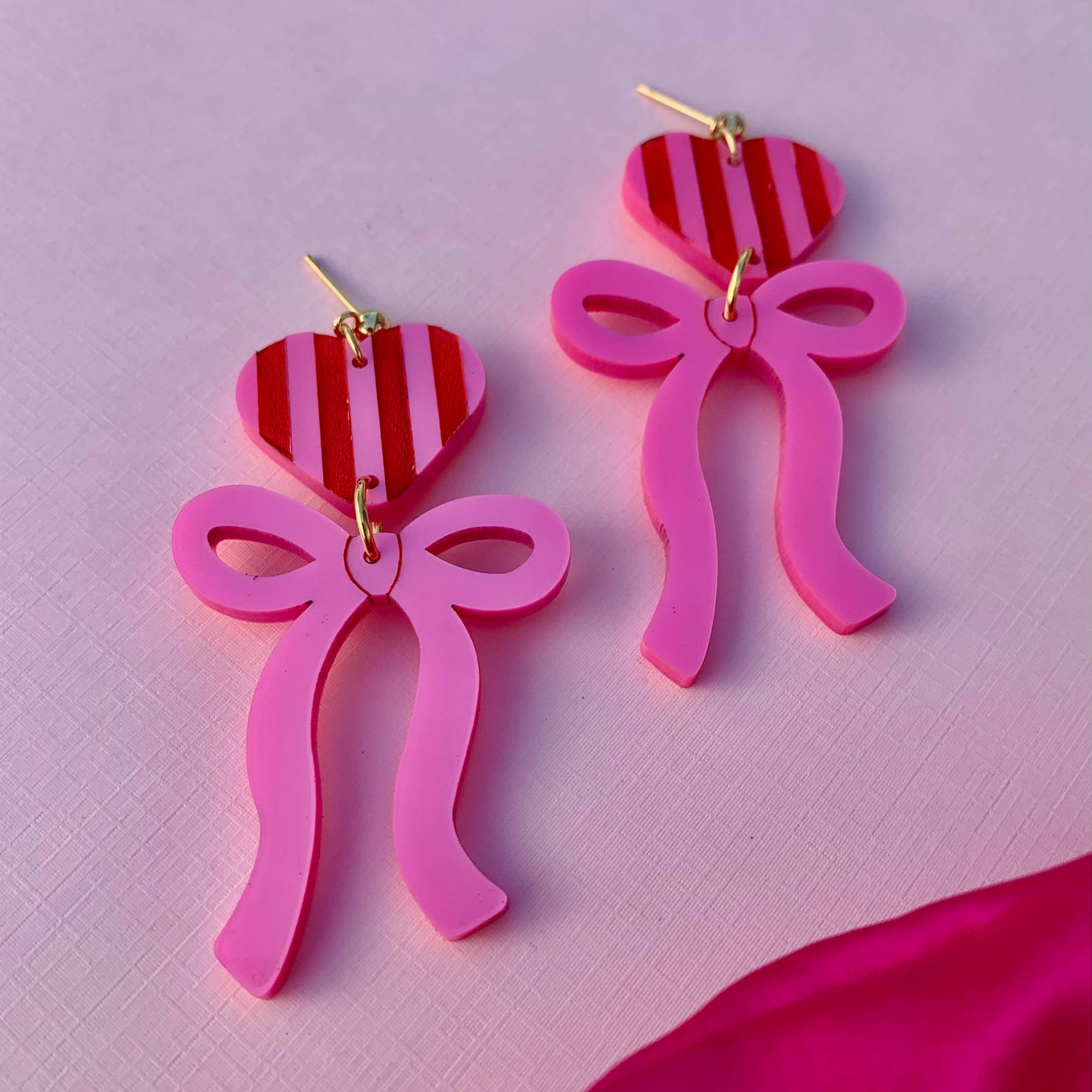 Pink bow-shaped earrings with red and white striped hearts on a pink background