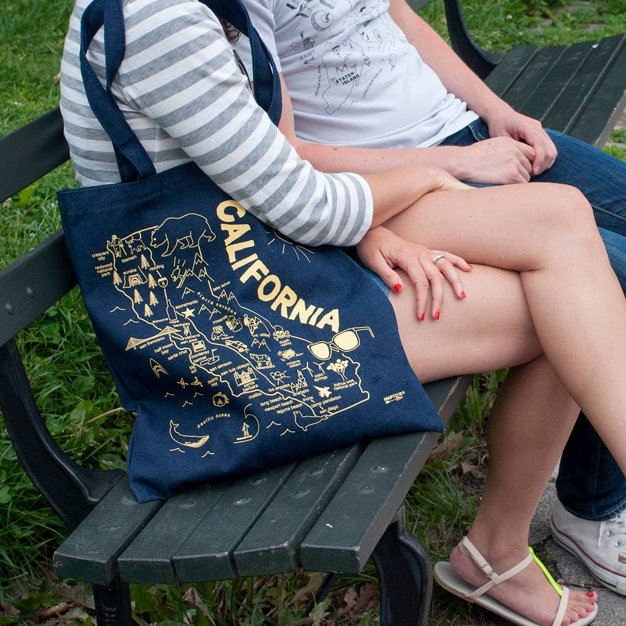 Two people sitting on a bench with a blue tote bag featuring 'California' text.