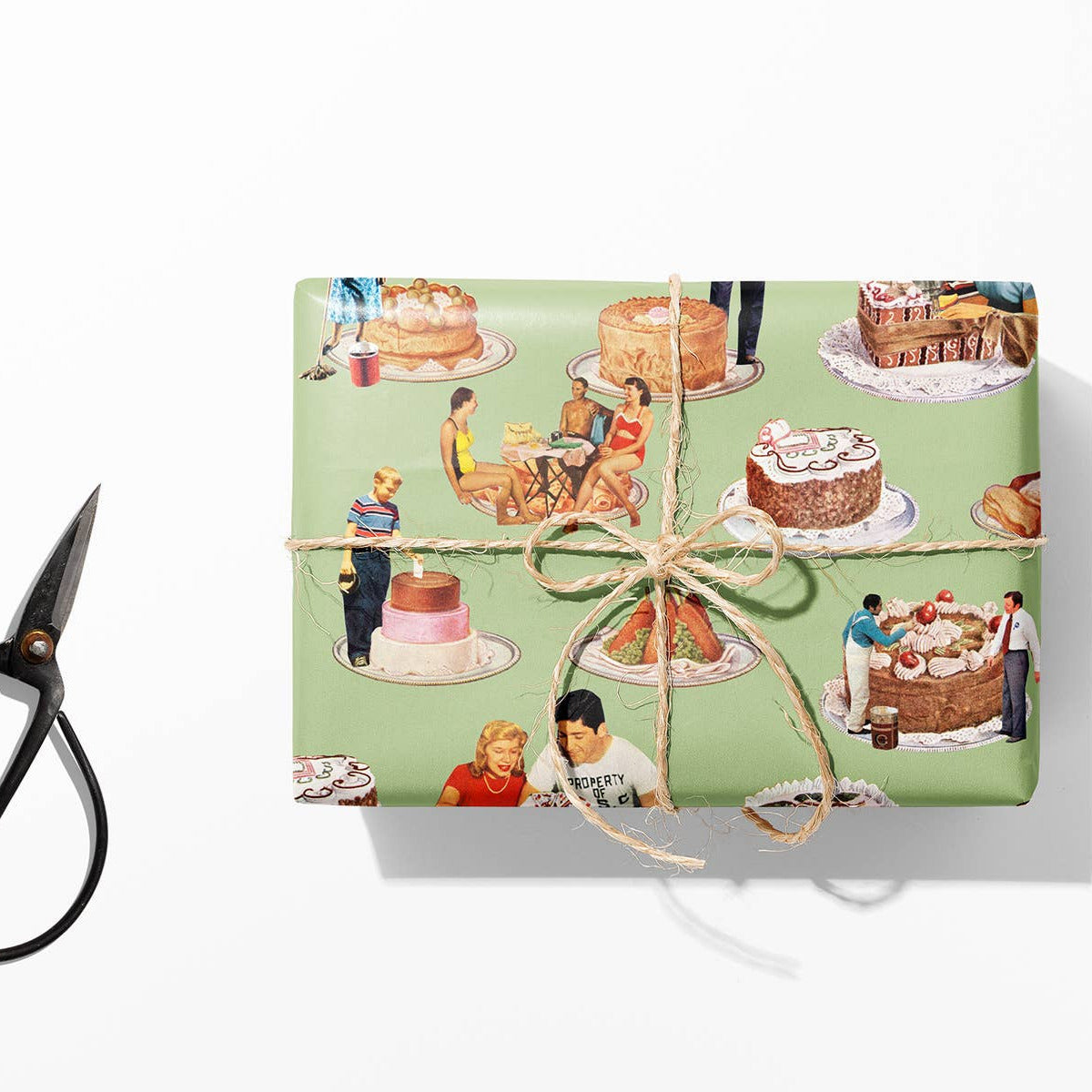 Gift box wrapped in paper with a vintage-style illustration of people and cakes, tied with string, next to scissors and twine on a white background.