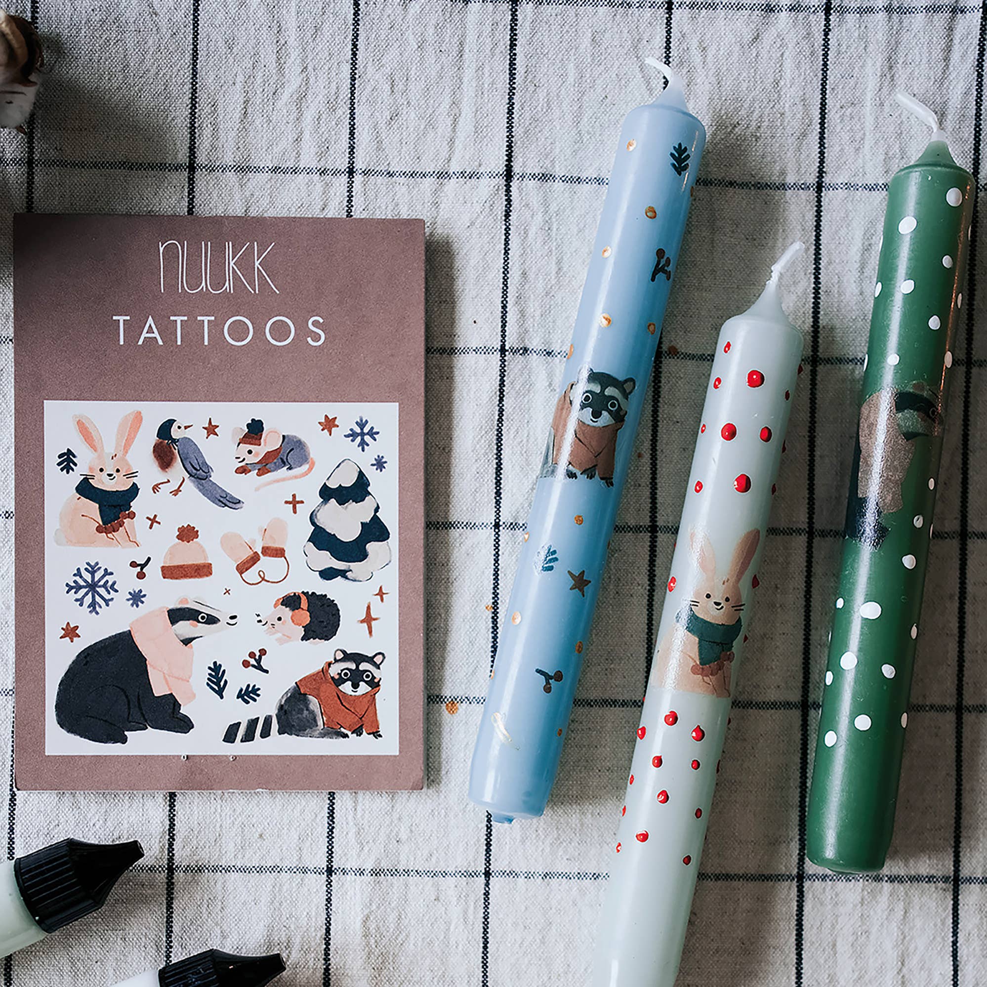 A flat-lay of tattoo ink sticks and a tattoo sheet on a black-and-white checkered cloth. The sheet reads 'NUKK TATTOOS' and shows winter animal illustrations including raccoons, rabbits, and snowflakes.