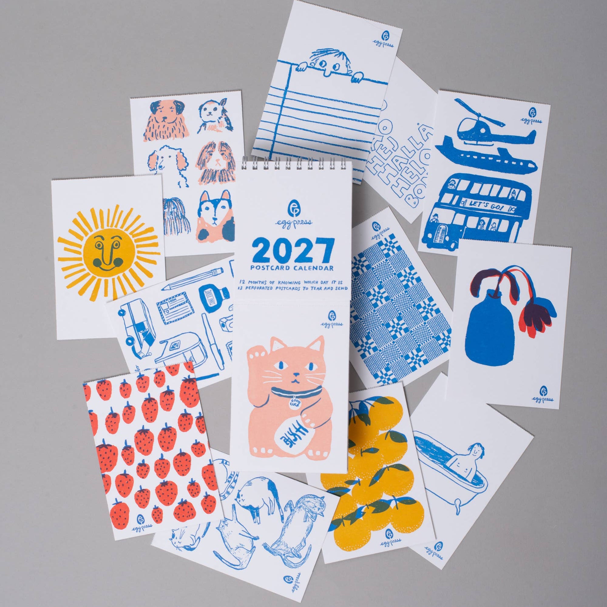 A flat-lay arrangement of multiple small printed cards and sheets featuring blue, red, yellow, and pink illustrations on a light gray background. The central sheet shows the year 2027 and a cat, while surrounding sheets include a sun, strawberries, a blue bag, a helicopter, and other simple graphic designs.
