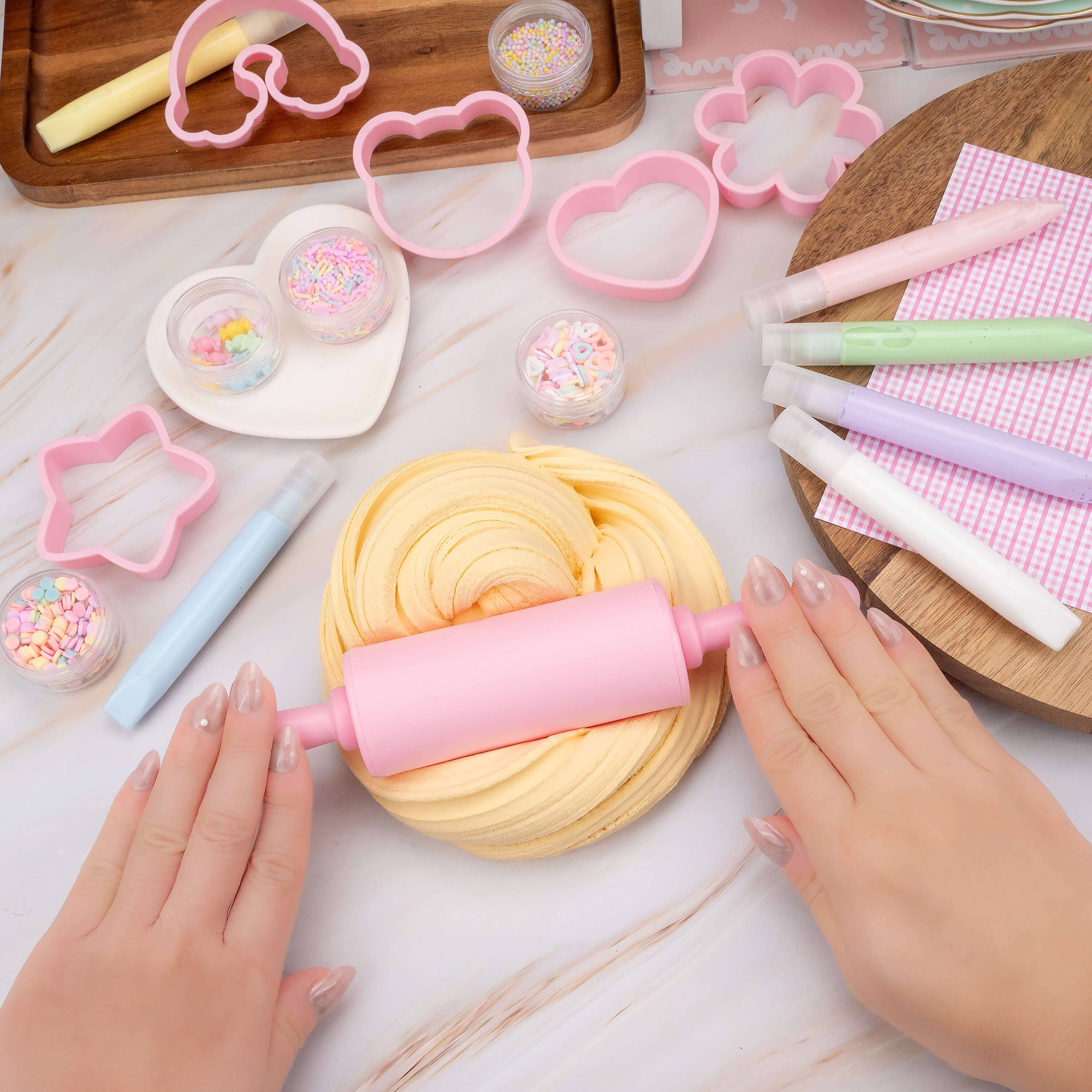 Hands using a pink rolling pin on a yellow dough with cookie cutters and sprinkles in the background.
