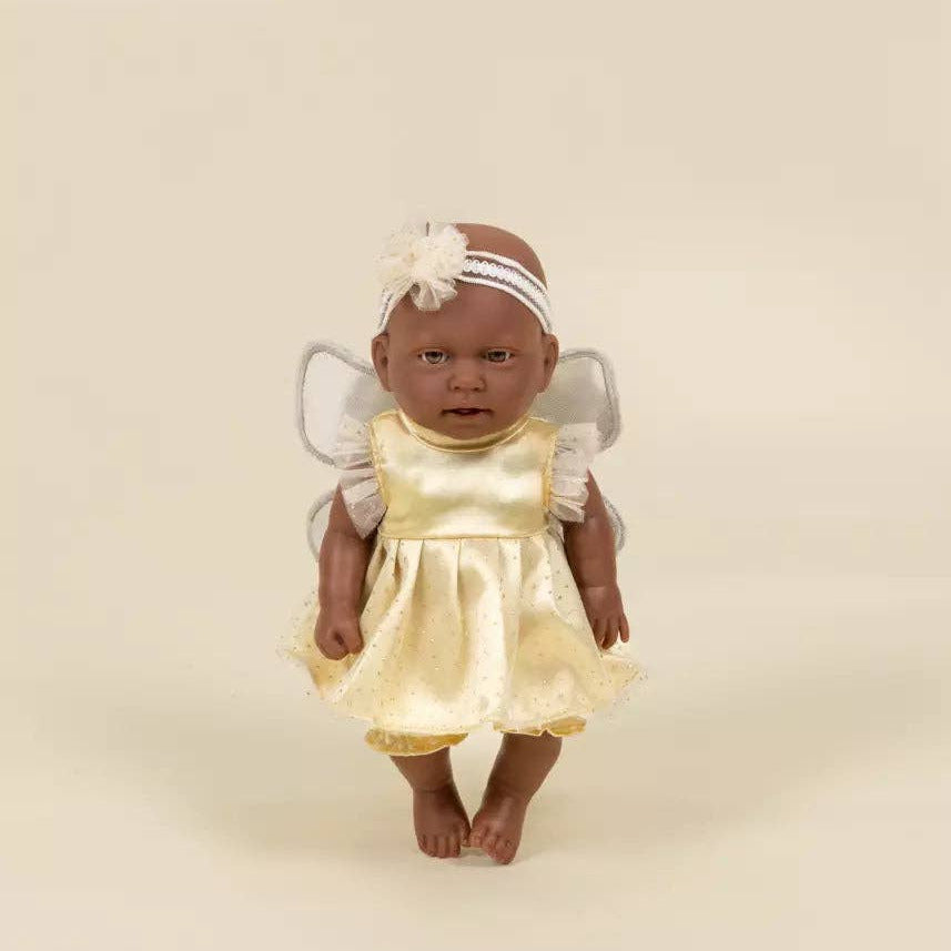 A baby doll with dark skin and a bald head, wearing a shiny pale yellow angel dress, a white floral headband, and small white wings, standing against a plain beige background.