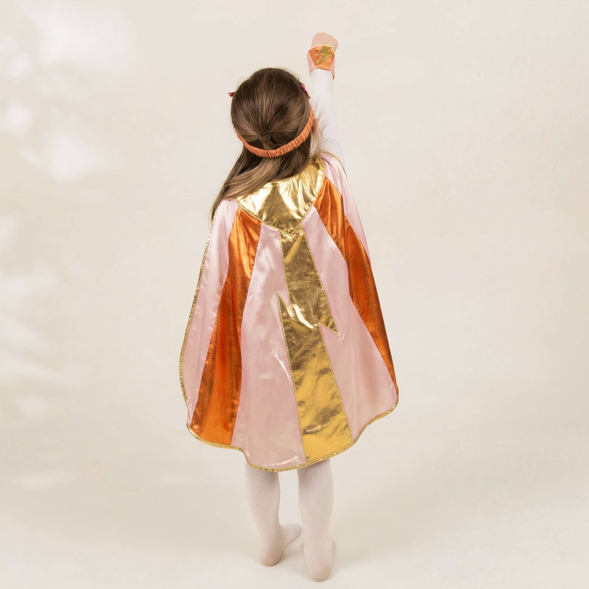 A child viewed from behind wearing a shiny costume cape with vertical panels of orange, gold, and pale pink, along with white tights and a matching headband, against a plain light background.