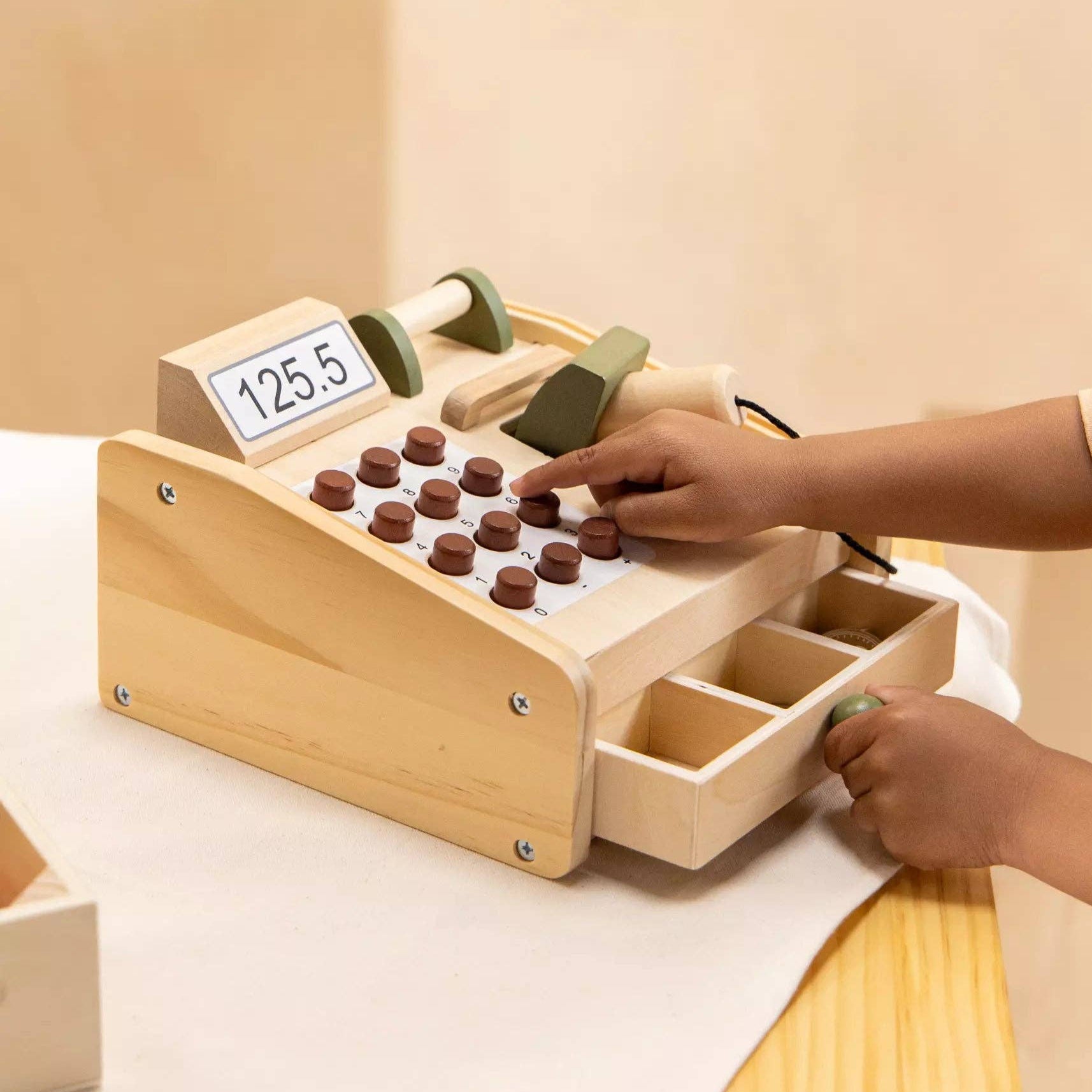 Child playing with a wooden toy cash register on a light surface.