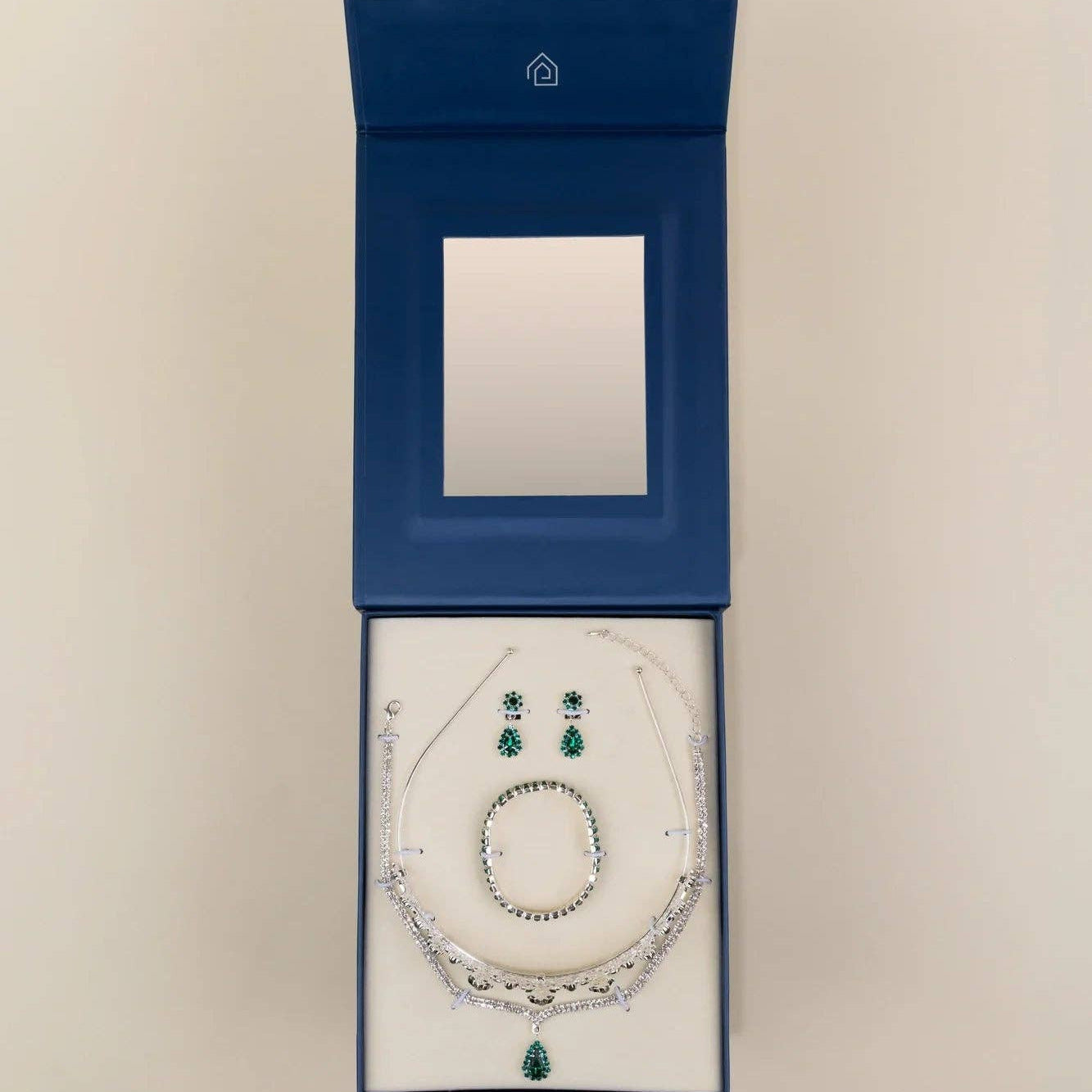A jewelry set displayed inside an open dark blue presentation box on a light beige background. The set includes a silver-toned necklace with clear stones and a green teardrop pendant, matching drop earrings, and a round ring.