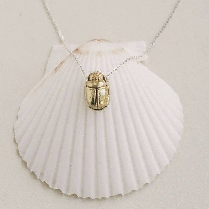 A gold beetle pendant necklace on a delicate silver chain, displayed on a white scallop shell against a light beige background.