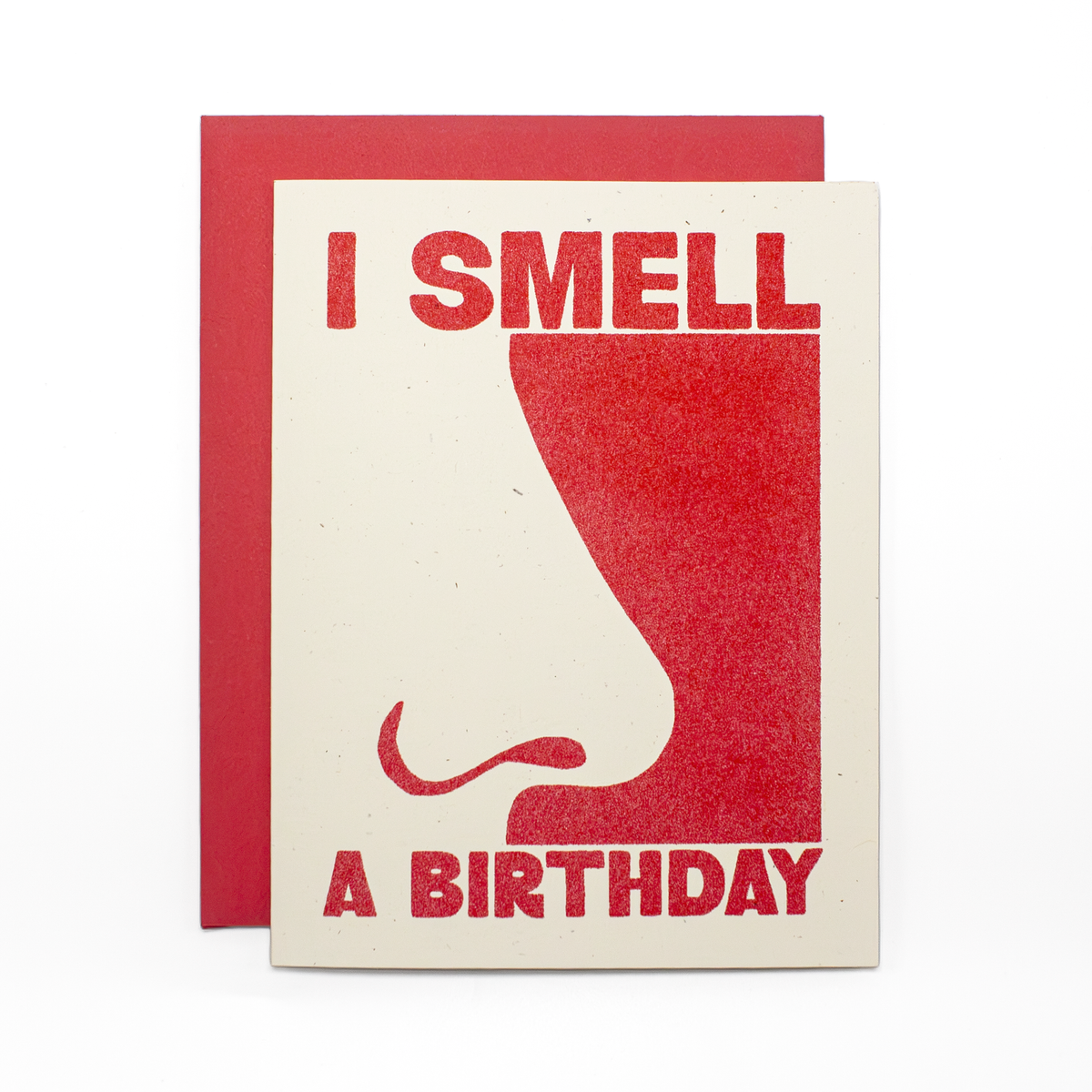 A square greeting card with a red envelope behind it. The card has a cream background with a large red graphic of a nose and the text 'I SMELL A BIRTHDAY' in red.