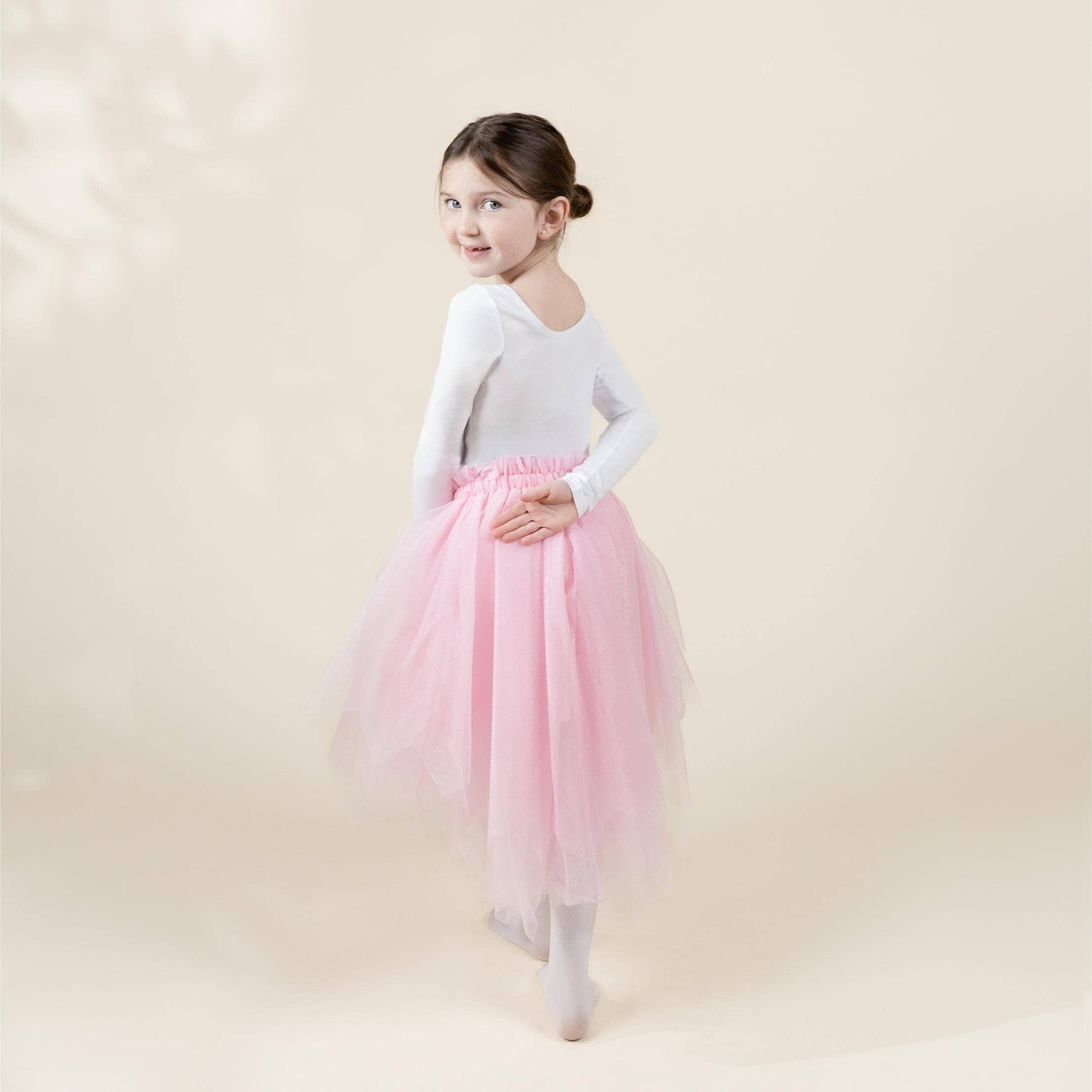 A young girl dancer wearing a white long-sleeve leotard and a fluffy light pink tutu skirt, standing in a ballet pose against a soft beige studio background.