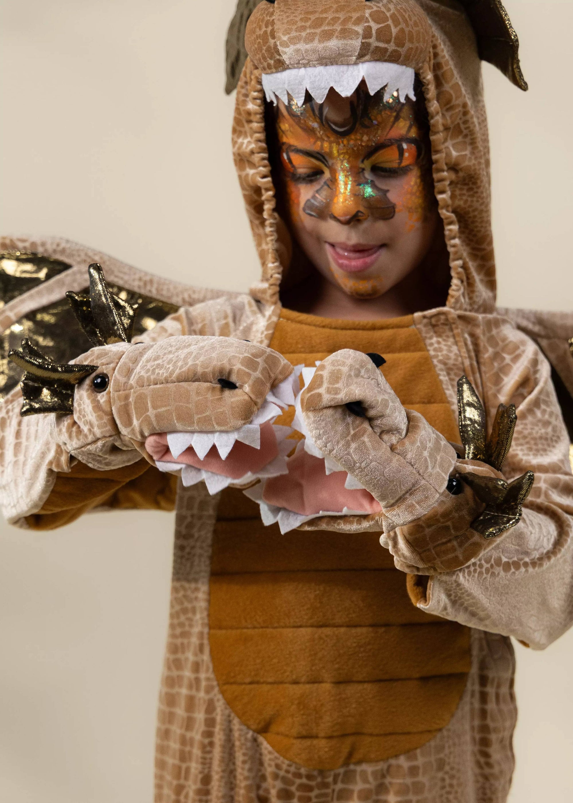 A child wearing a detailed dinosaur costume with a hood, textured brown and tan body, gold face paint, and matching dinosaur gloves with white teeth.