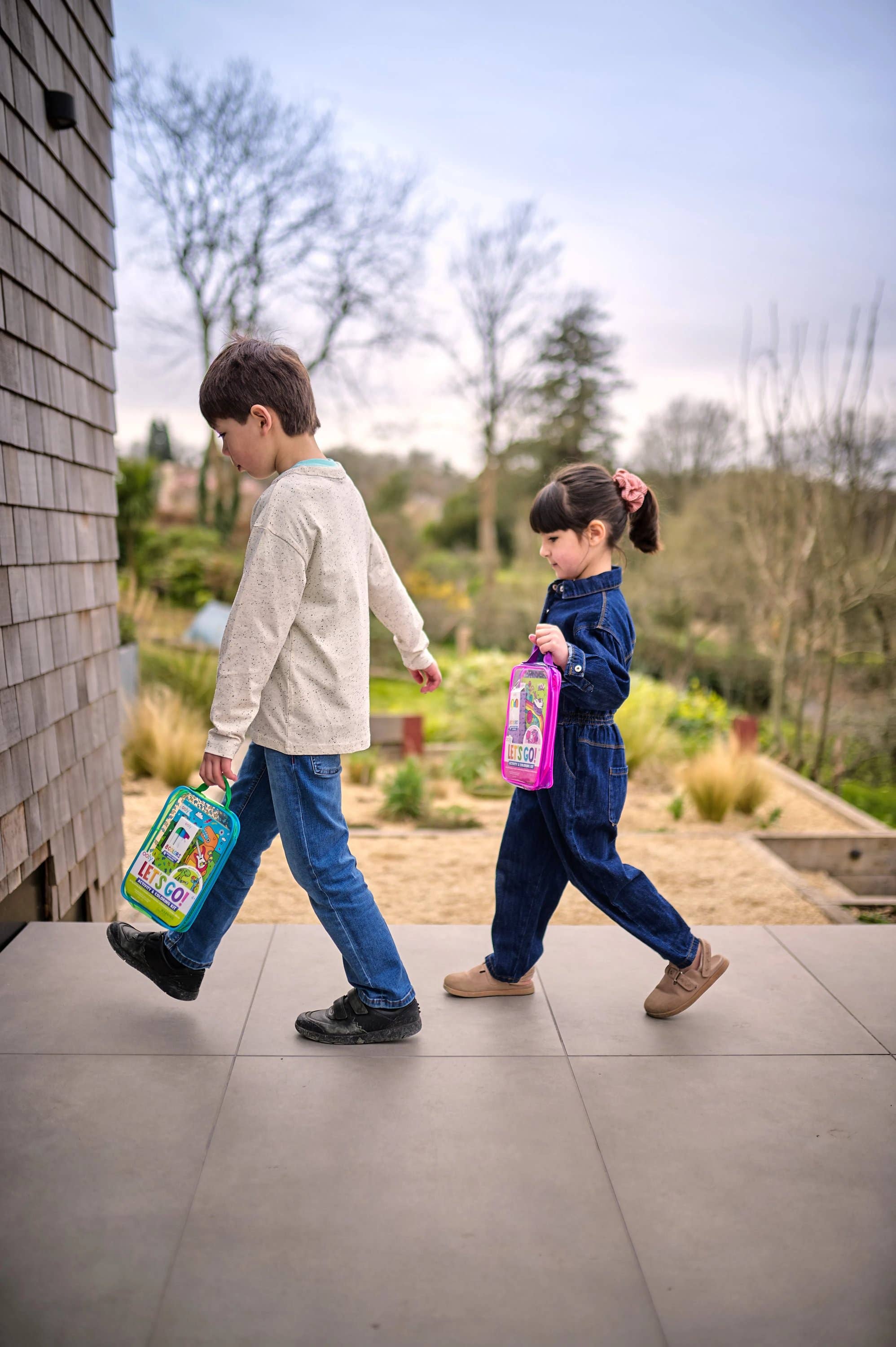 Two children walking outdoors with toys, one holding a colorful book and the other a pink toy.