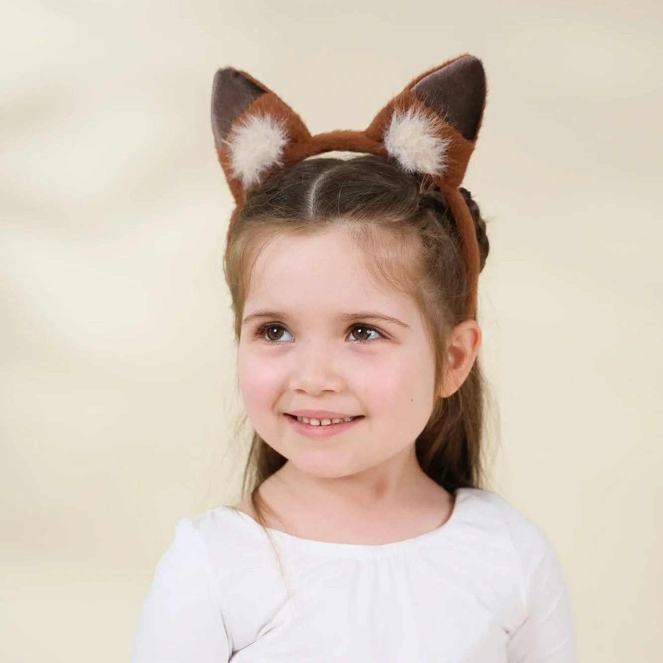 A young girl with brown hair wearing a brown and white faux fur animal ears headband, smiling against a soft beige background.
