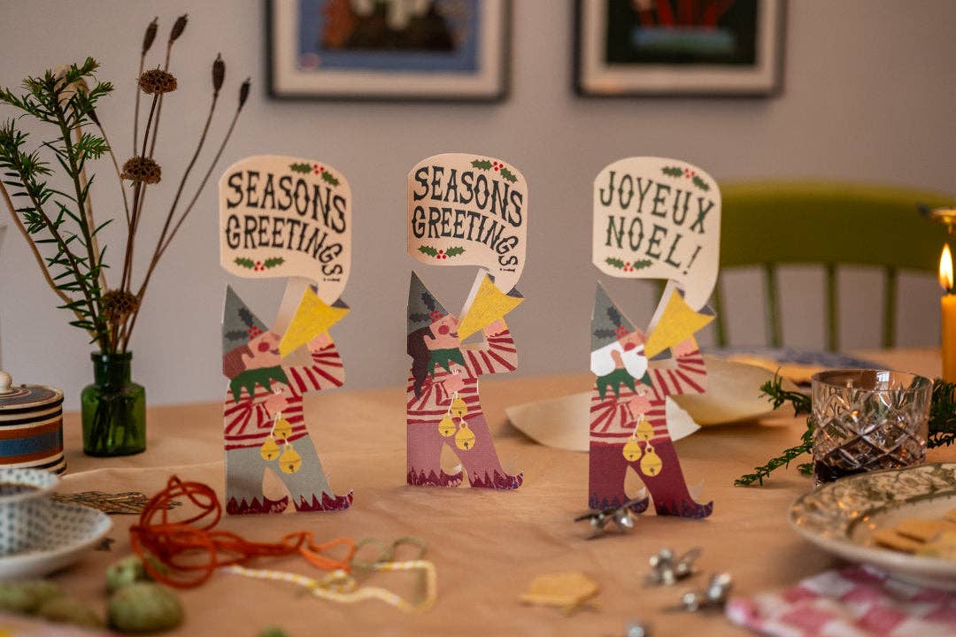 Three handmade paper holiday table decorations shaped like Santa Claus stand on a wooden dining table, with two reading 'SEASONS GREETINGS' and one reading 'JOYEUX NOEL!'. The scene includes a green bottle vase with pine branches, holiday ornaments, plates, and a candle in a real indoor setting.