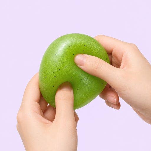 A pair of hands squeezing a green kiwi-shaped stress toy against a solid light purple background.