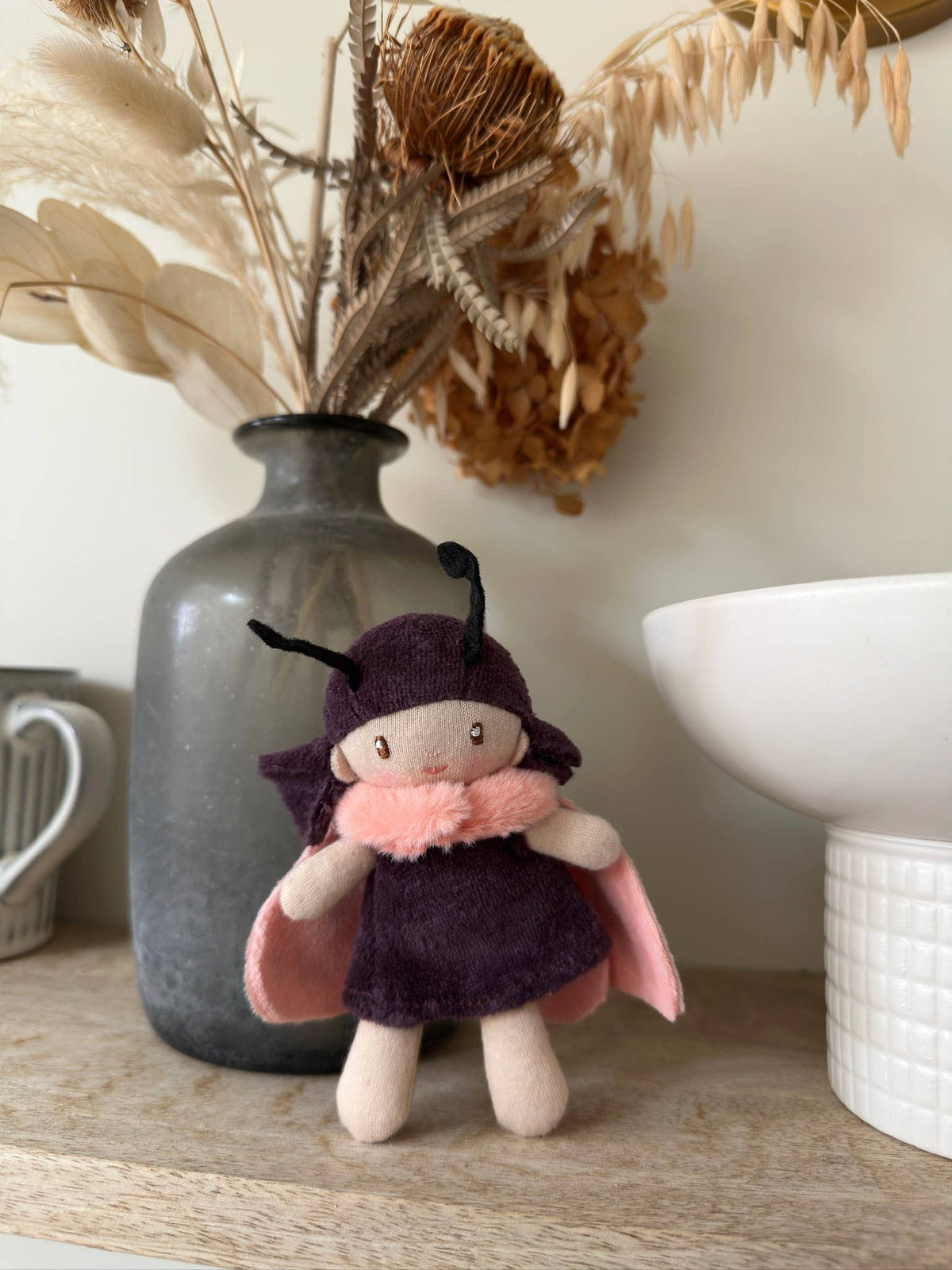 Small plush toy with a purple outfit and pink cape on a wooden surface with decorative elements.