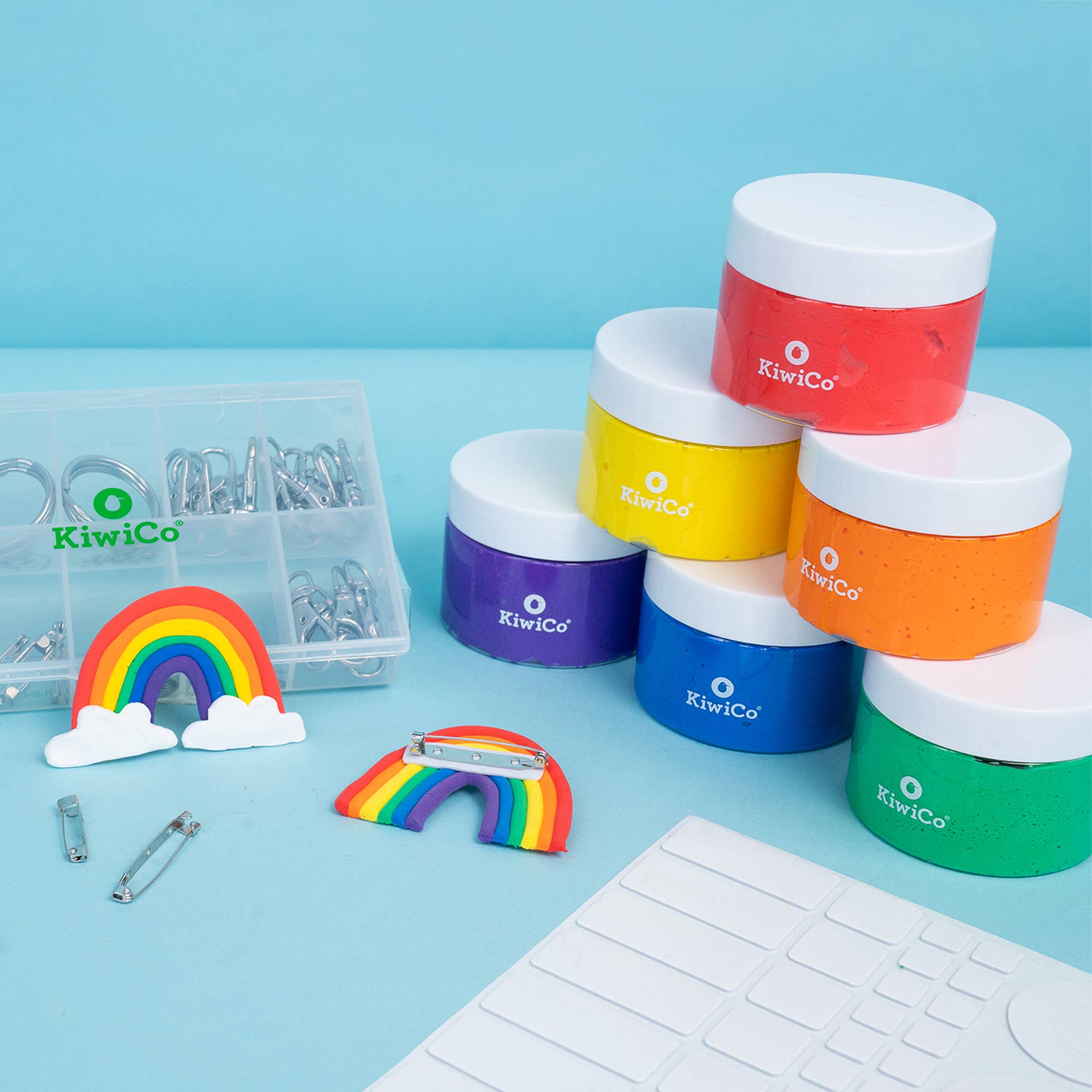A set of KiwiCo branded hair pins and hair clips displayed on a light blue surface. The set includes a clear organizer box with silver metal clips, a rainbow-shaped pin with white cloud accents, and a stack of five colorful hair pins in red, yellow, orange, purple, and green with white lids.