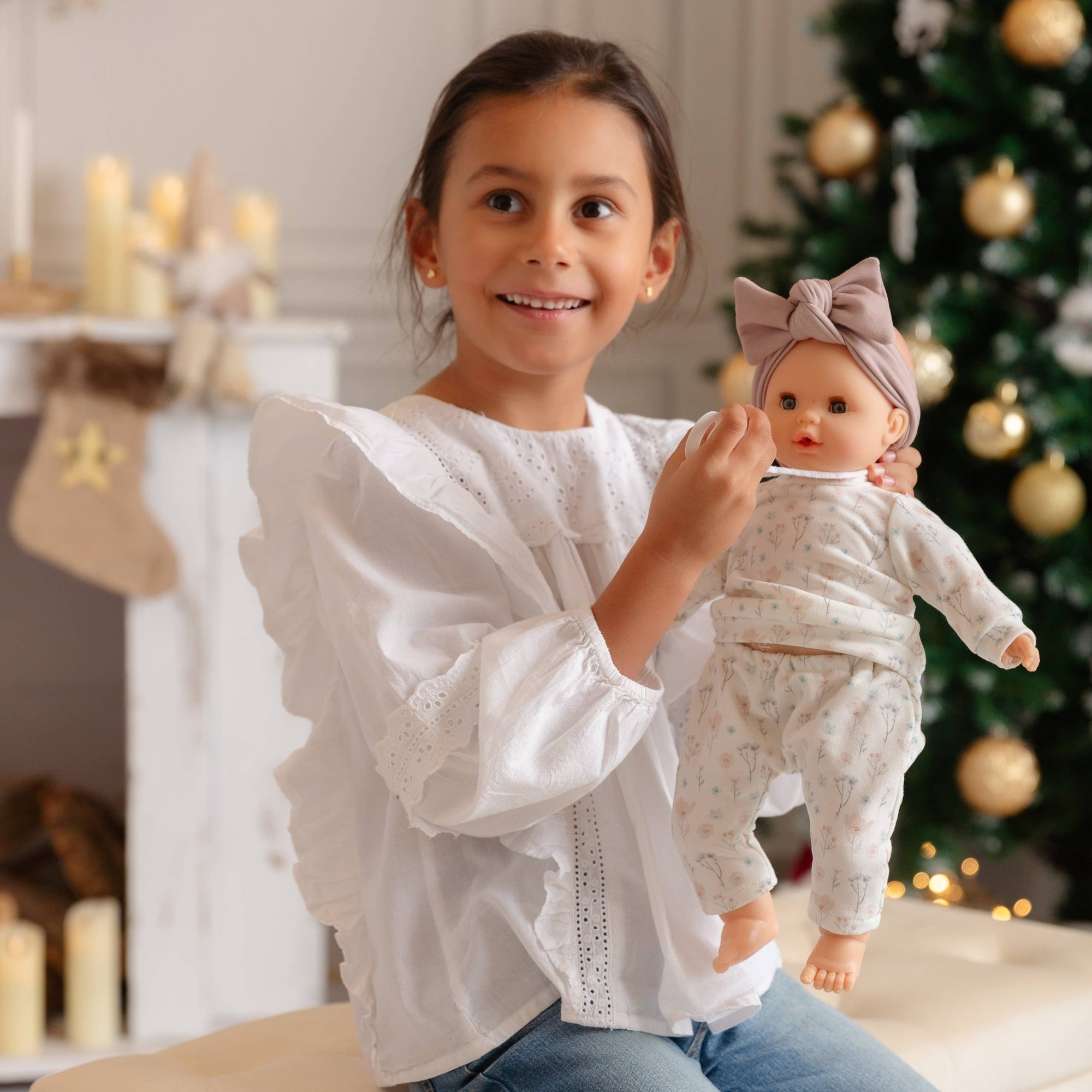 Young girl holding a baby doll in front of a decorated Christmas tree.