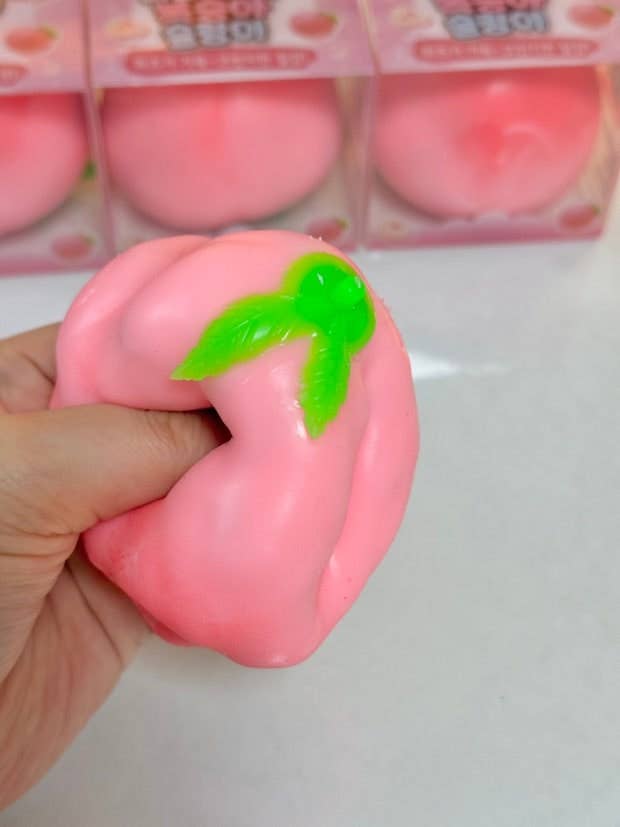A hand holding a pink peach-shaped squishy toy with a green stem, with packaged peach toys blurred in the background.
