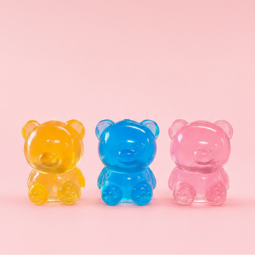 Three translucent teddy bear-shaped gummy candies in yellow, blue, and pink, arranged side by side on a solid pink background.