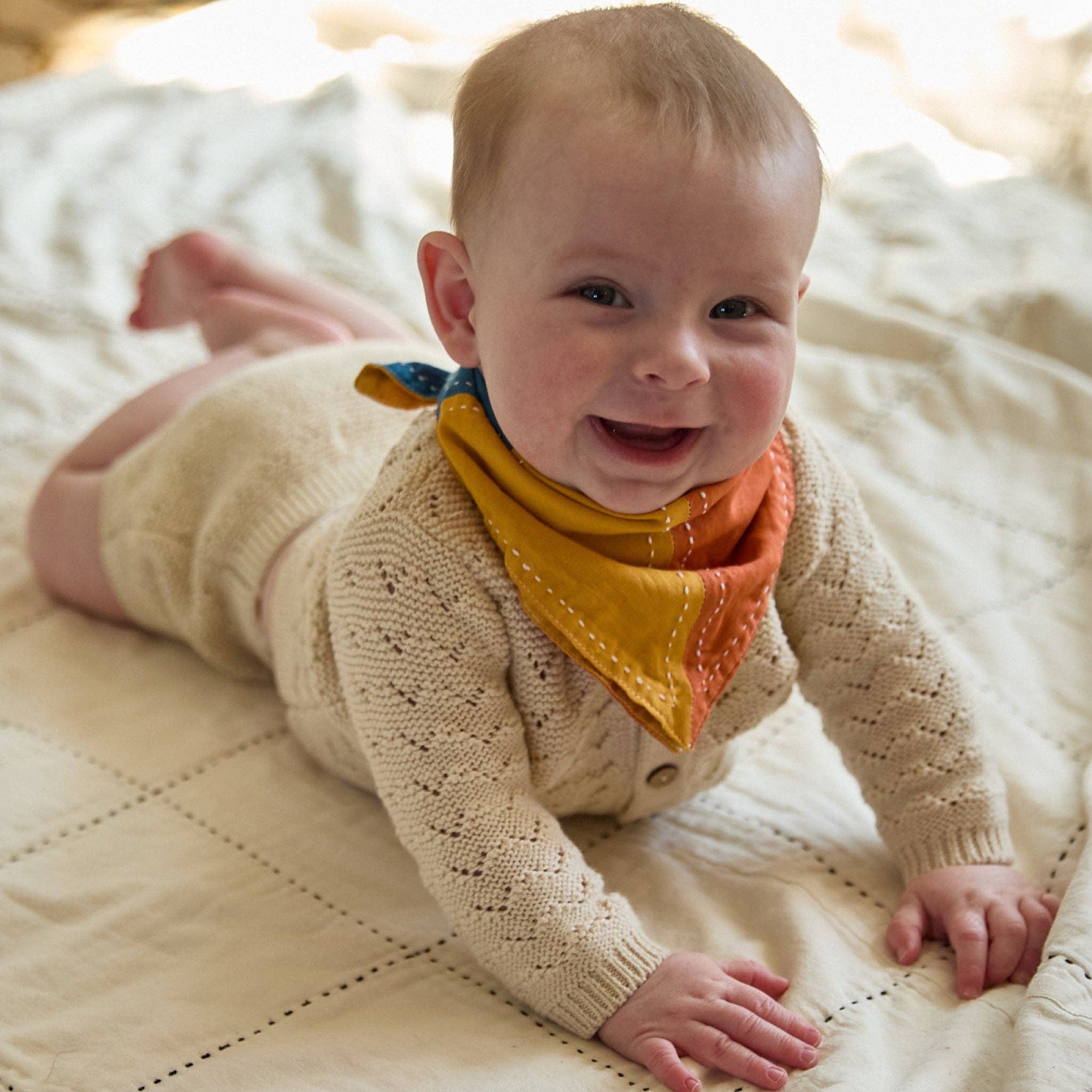 Baby wearing a beige knitted sweater and colorful bandana lying on a quilted surface.