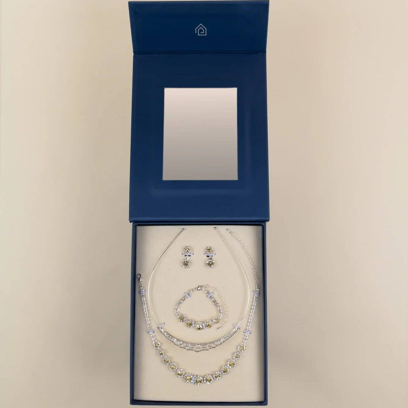 A jewelry set displayed inside an open dark blue presentation box on a light beige background. The set includes a necklace, bracelet, and matching earrings with clear rhinestone-like embellishments on silver-toned chains.