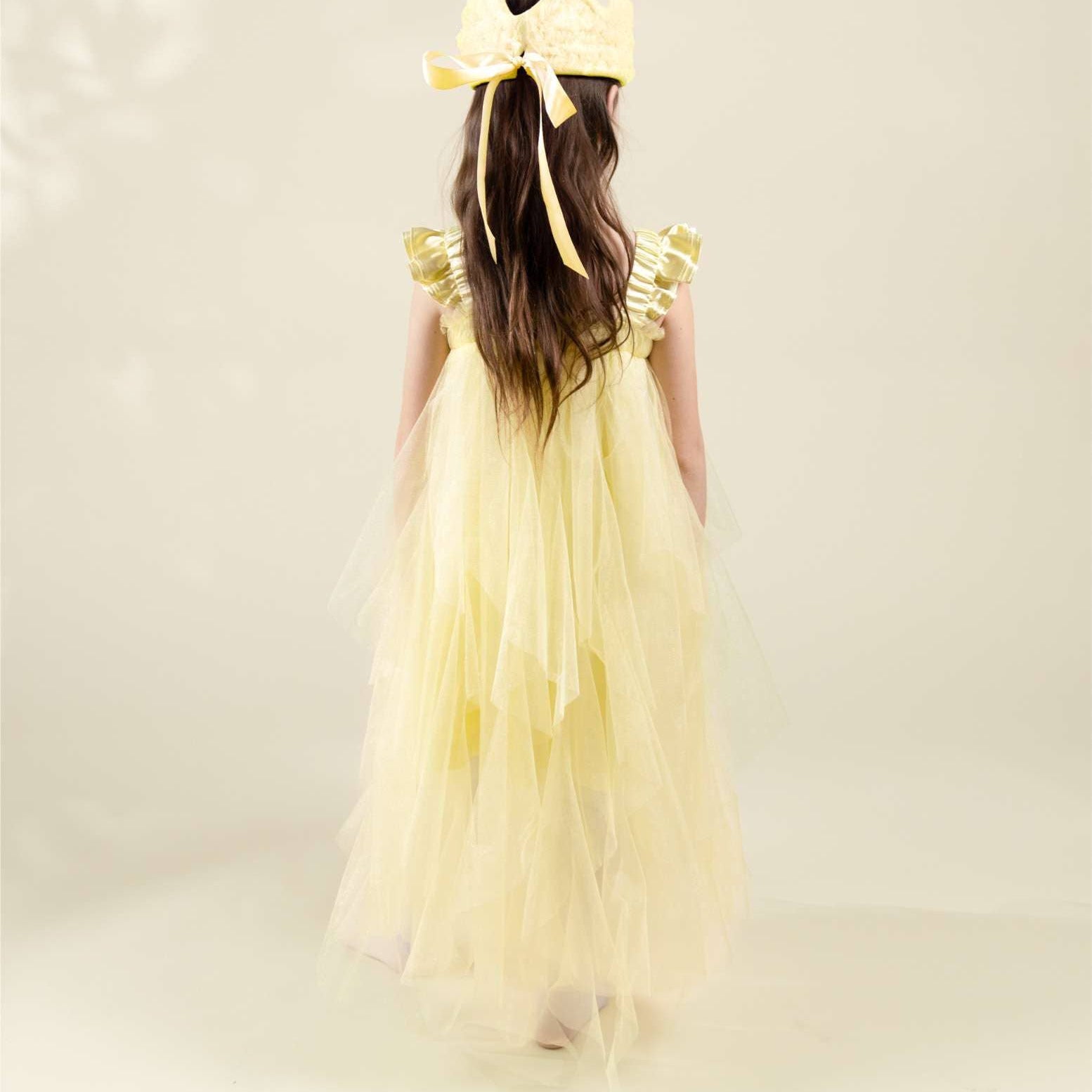 Young girl wearing a yellow dress with a large bow on her head against a light background
