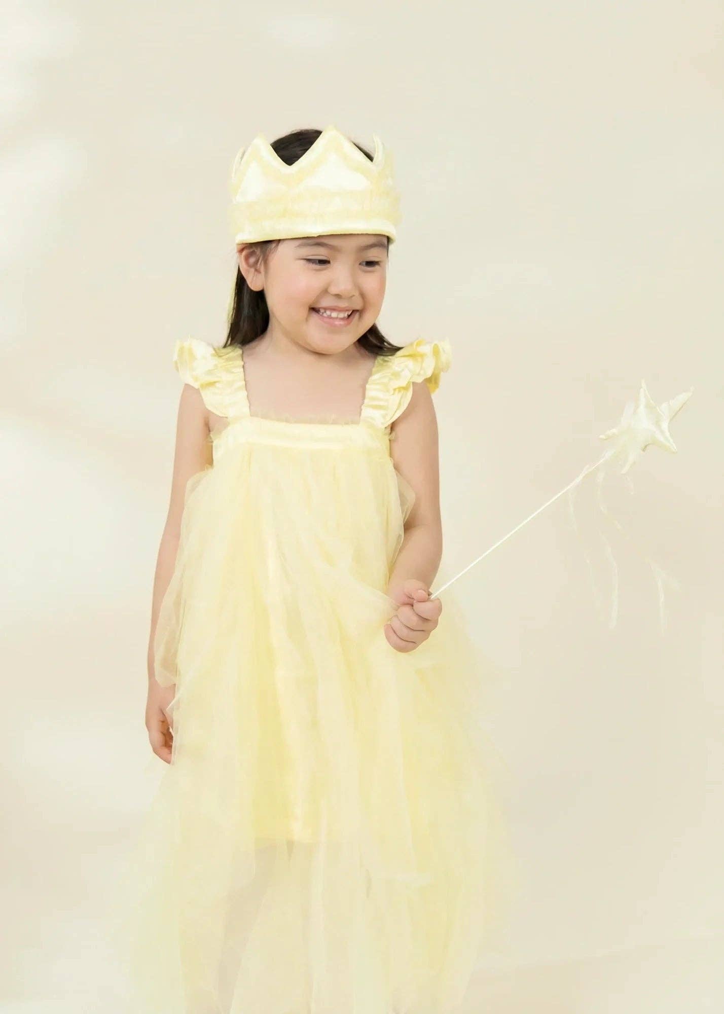 A young girl wearing a light yellow princess costume with a matching crown and holding a star wand, set against a soft, warm beige background.