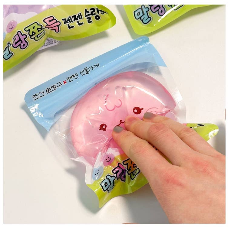 A hand pressing a pink cat-shaped squishy toy inside a clear plastic bag with a blue header, placed on a white surface with other colorful product packages partially visible.