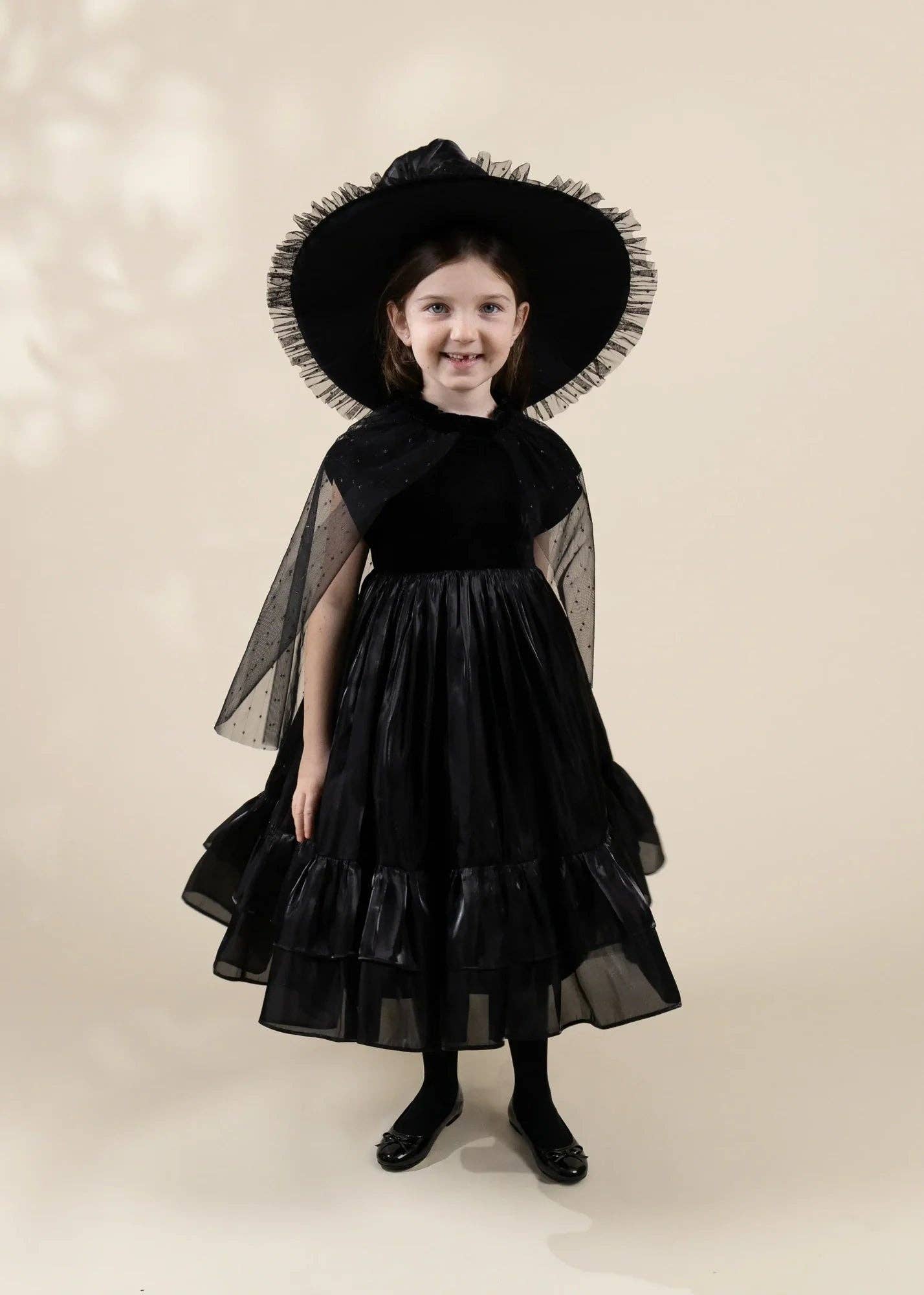 A young girl modeling a black witch costume consisting of a tiered tulle dress, a sheer black cape, and a matching wide-brimmed black hat with ruffled trim, set against a solid beige studio background.