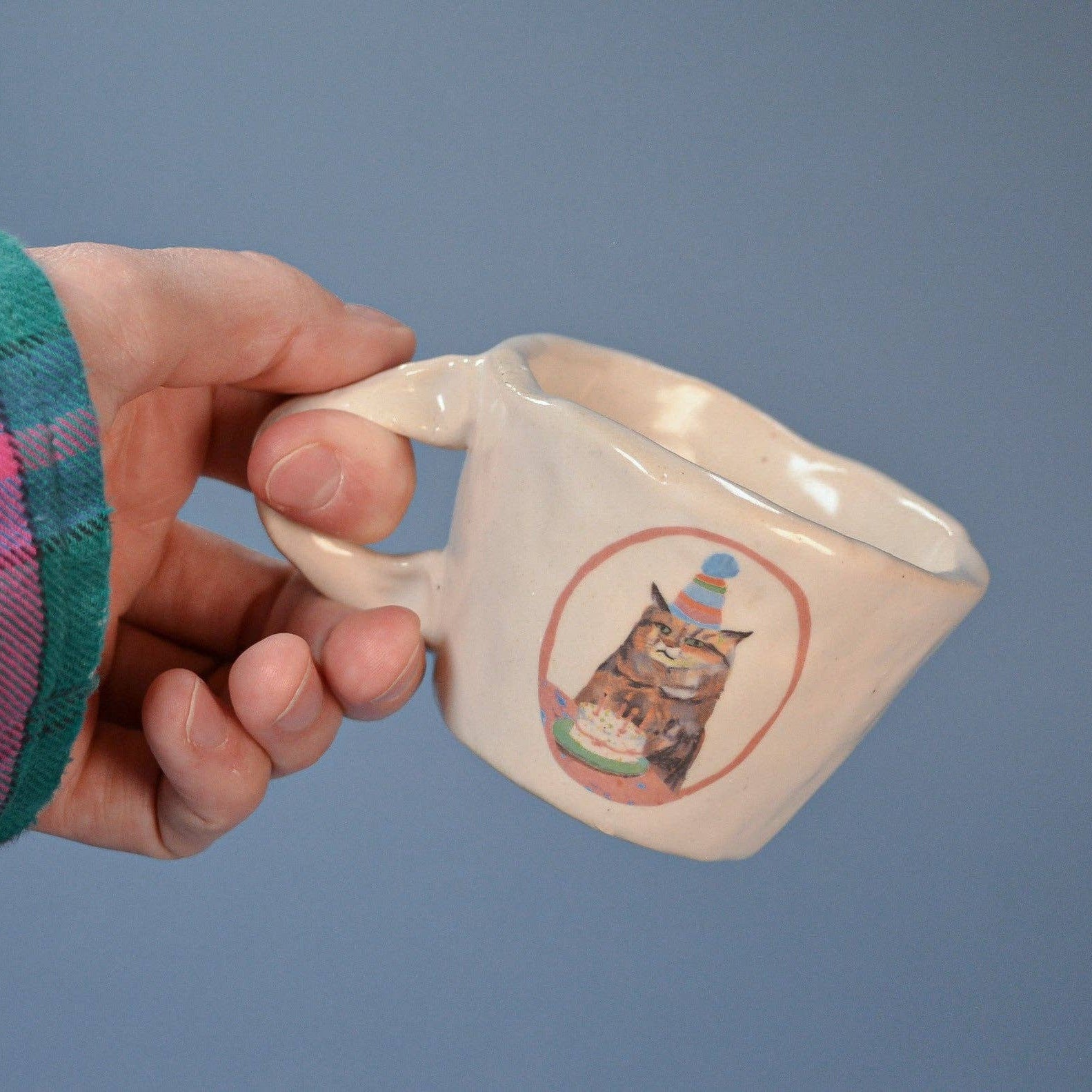 Hand holding a white mug with a cat illustration on a blue background