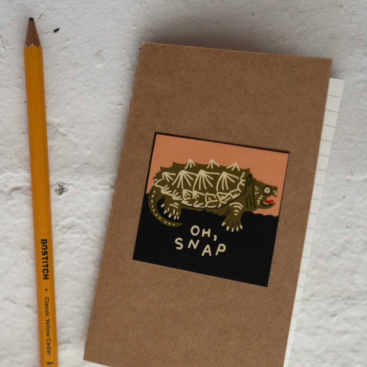 Brown notebook with a dinosaur illustration and 'OH SNAP' text, next to a yellow pencil on a textured surface.