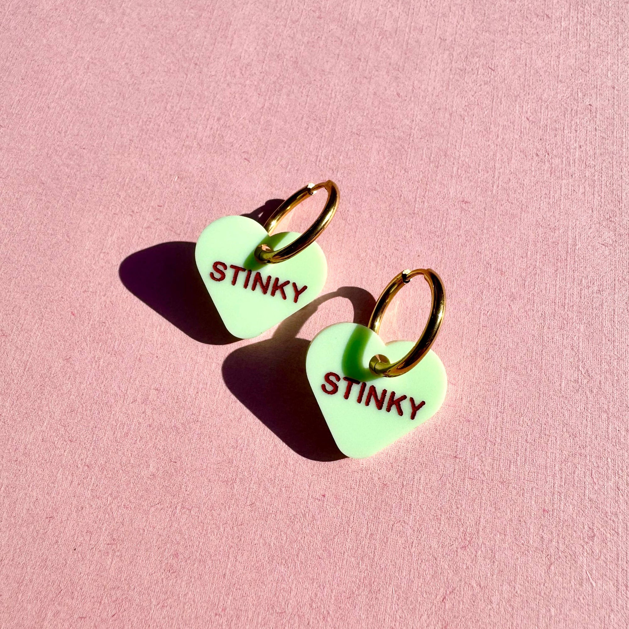 Heart-shaped earrings with 'STINKY' text on a pink background