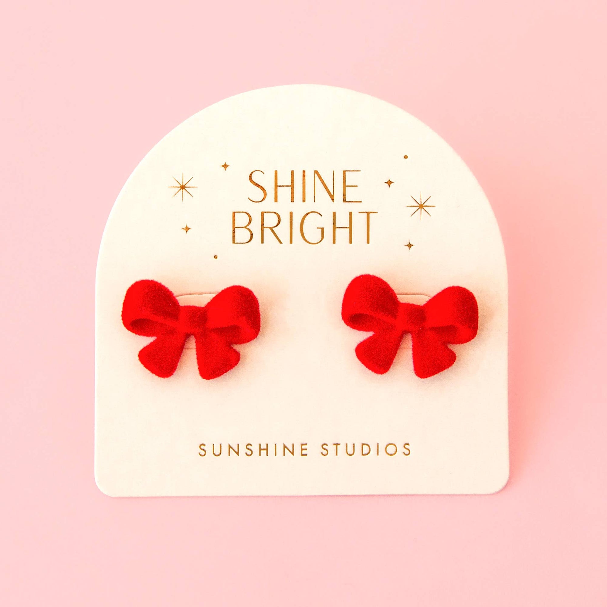 A pair of red bow stud earrings displayed on a white arched backing card that reads 'SHINE BRIGHT' and 'SUNSHINE STUDIOS' against a soft pink background.
