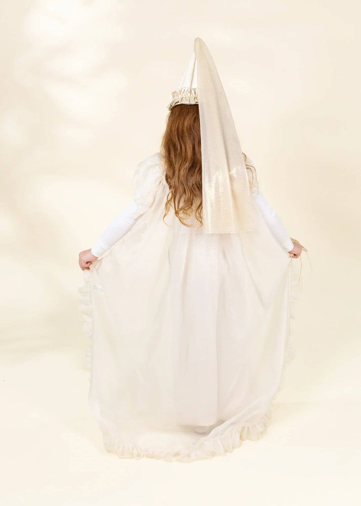A child wearing a white nun habit and veil, photographed from behind against a warm cream studio background.