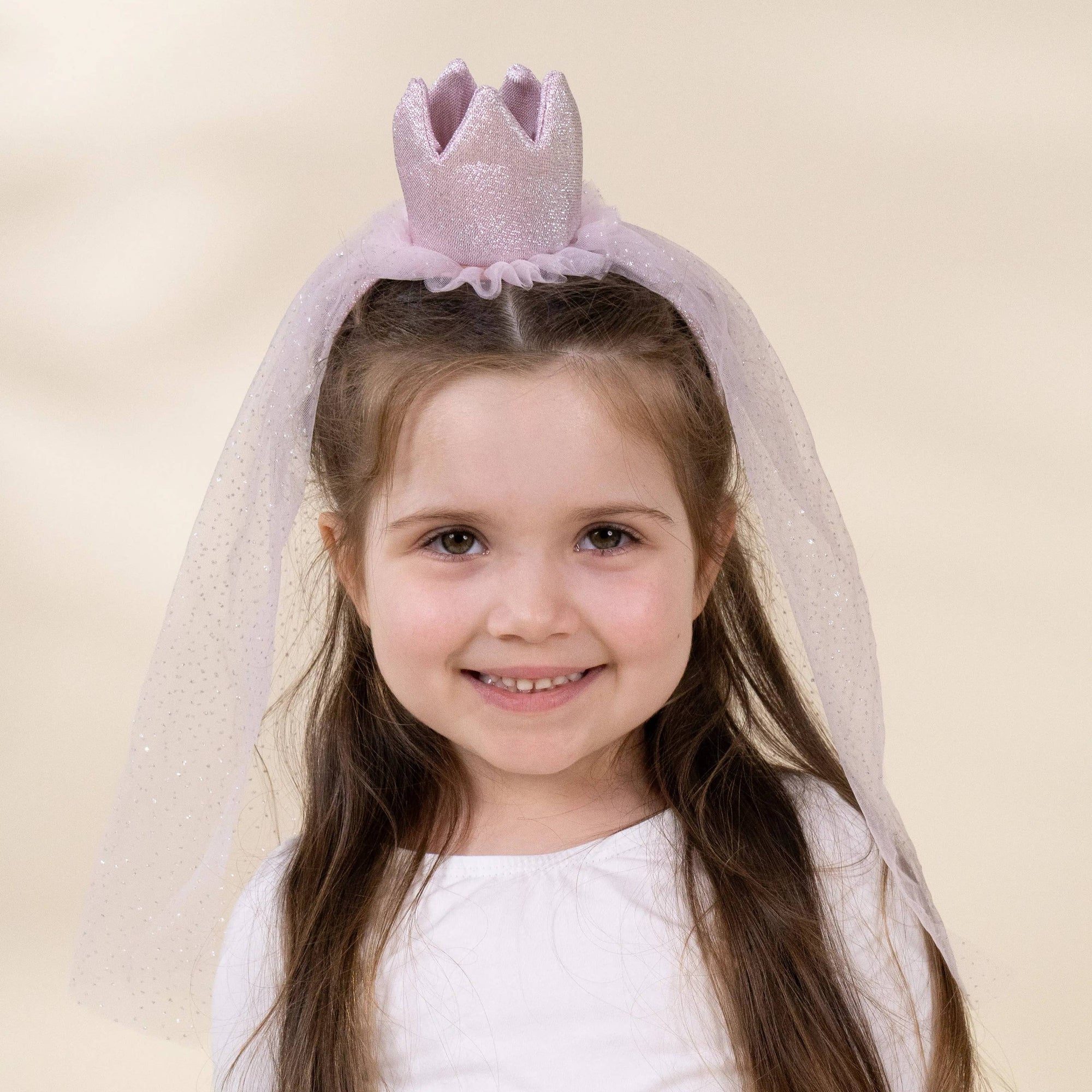 A young girl with long brown hair wearing a light pink glitter crown headband with a matching sheer tulle veil, posing against a soft beige studio background.