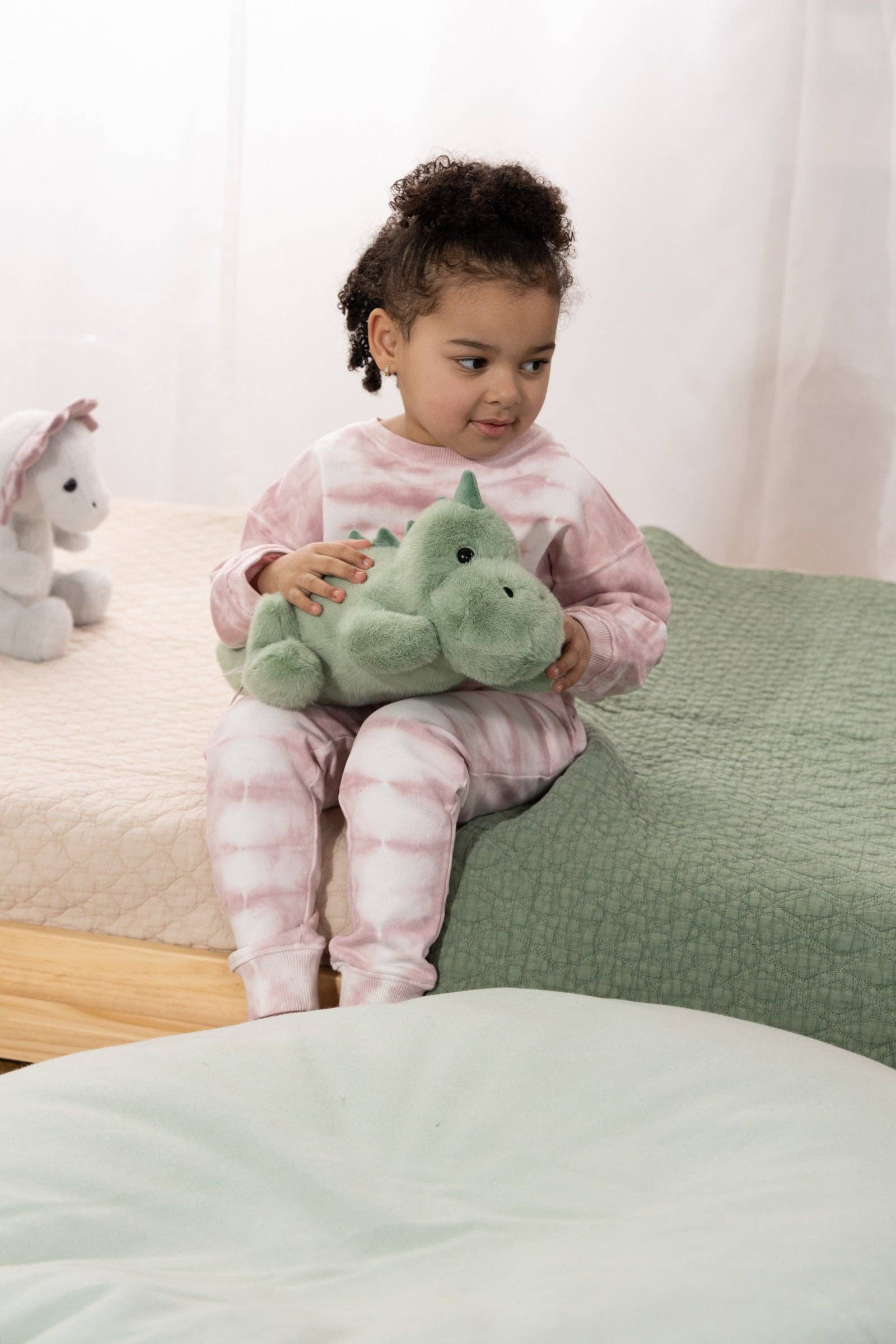 Child holding a green plush toy on a bed with a white curtain in the background