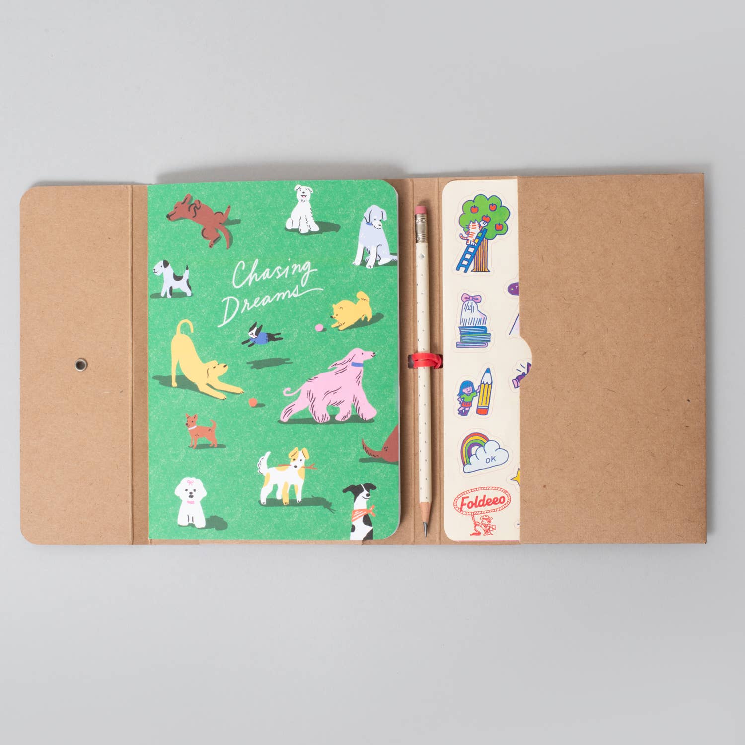 A folded kraft paper notebook or journal shown open on a light gray background. The left page is bright green with colorful dog illustrations and the words 'Chasing Dreams.