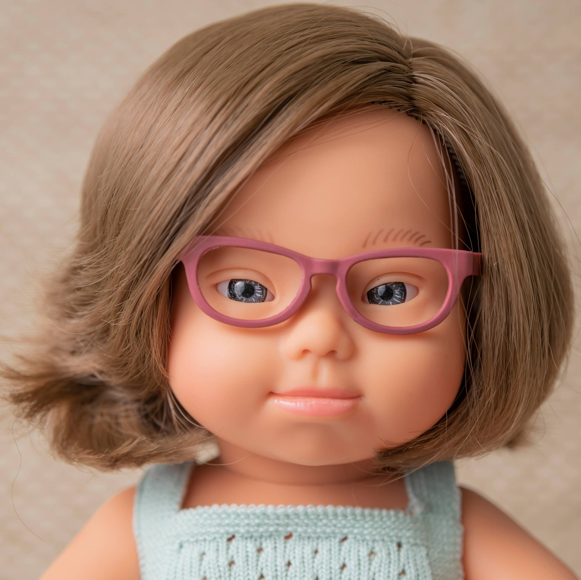 Doll with pink glasses and a light blue top against a beige background