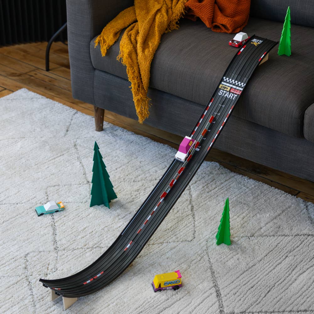 Toy race track with cars on a carpeted floor next to a sofa.