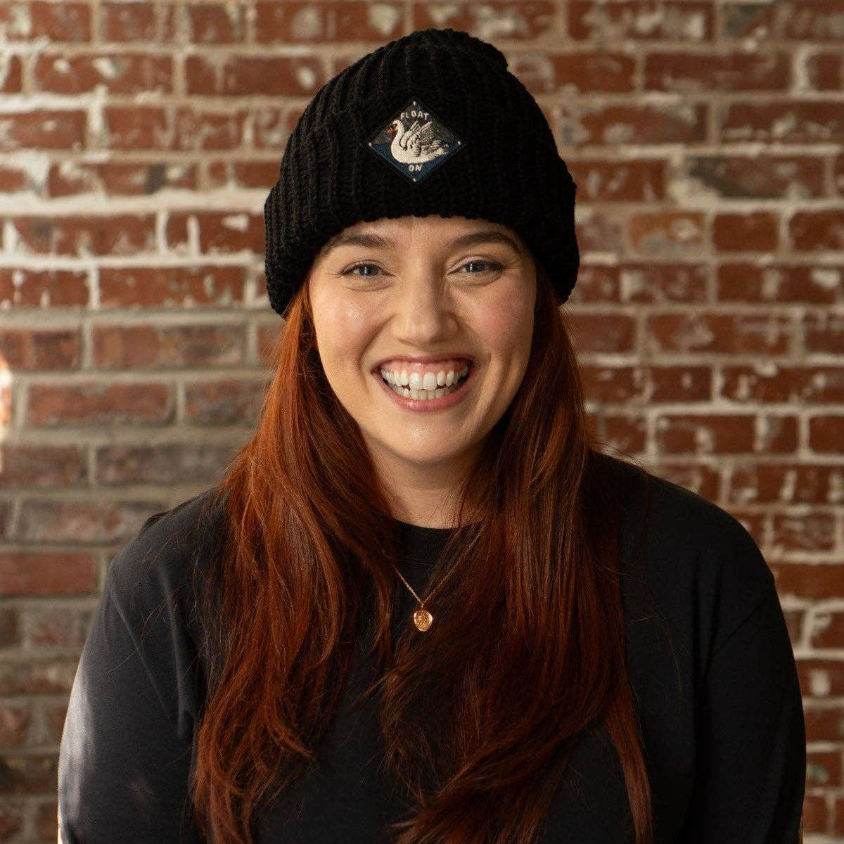 Person wearing a black beanie and dark clothing against a brick wall
